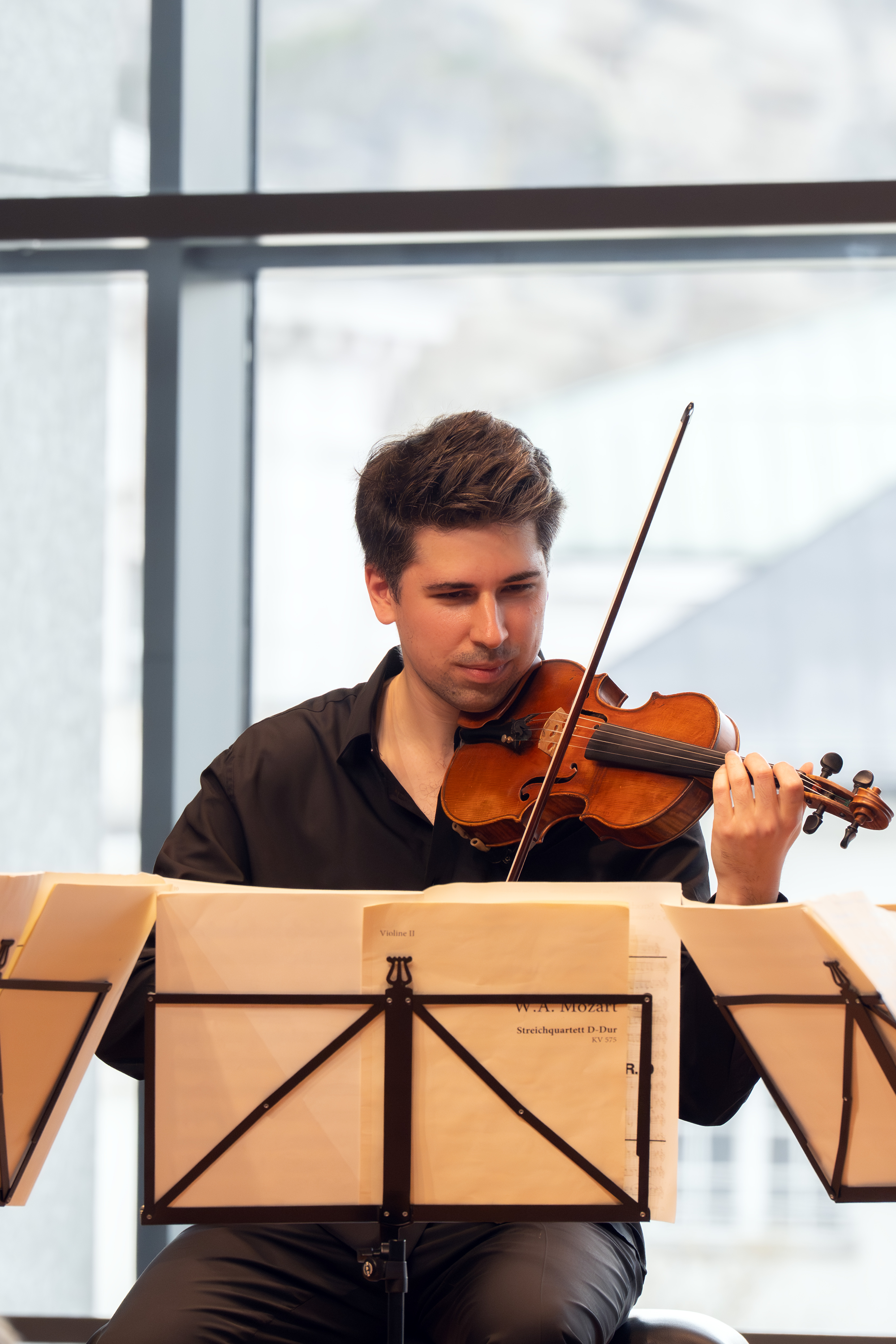 Ein Mann mit schwarzem Hemd spielt Geige, während er hinter Notenständern in einem hellen Raum mit großen Fenstern sitzt.