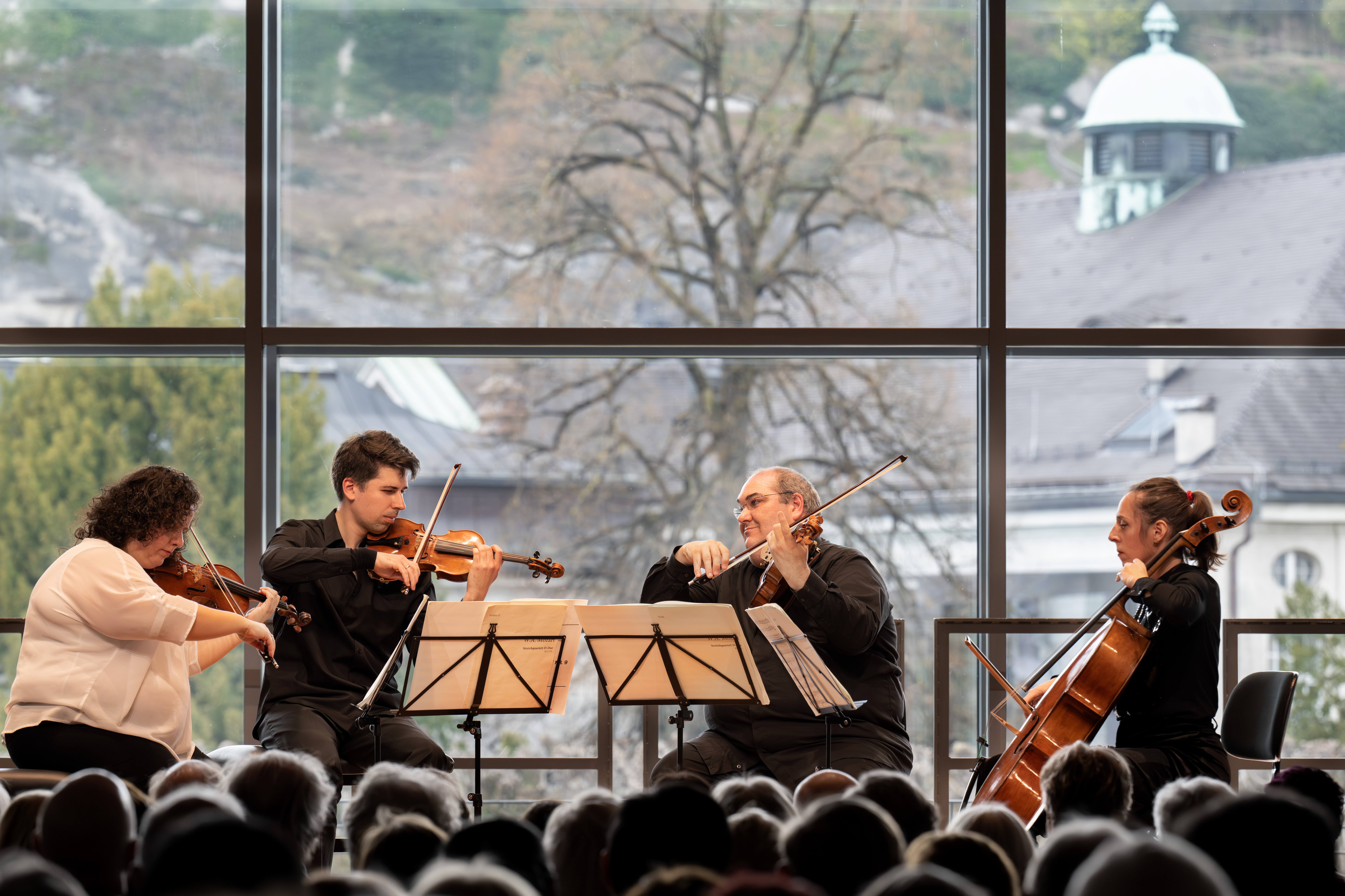 Ein Streichquartett spielt in einem Innenraum mit Notenblättern, umrahmt von großen Fenstern mit Bäumen und einem historischen Gebäude. Im Vordergrund sind die Zuhörer zu sehen.