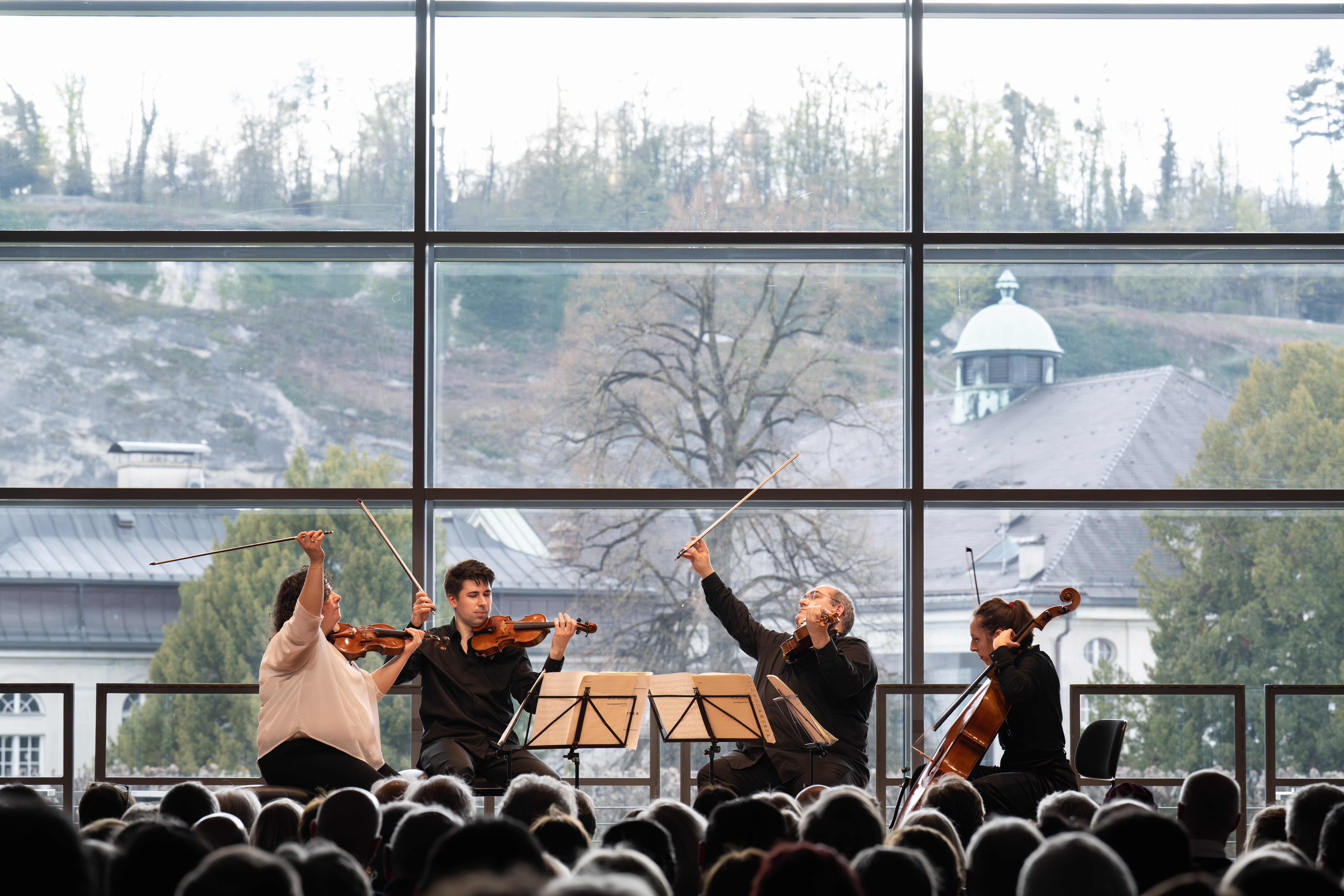 Ein Streichquartett spielt auf der Bühne vor einem großen Fenster mit Blick auf Bäume und Gebäude; ein Publikum beobachtet die Musiker.