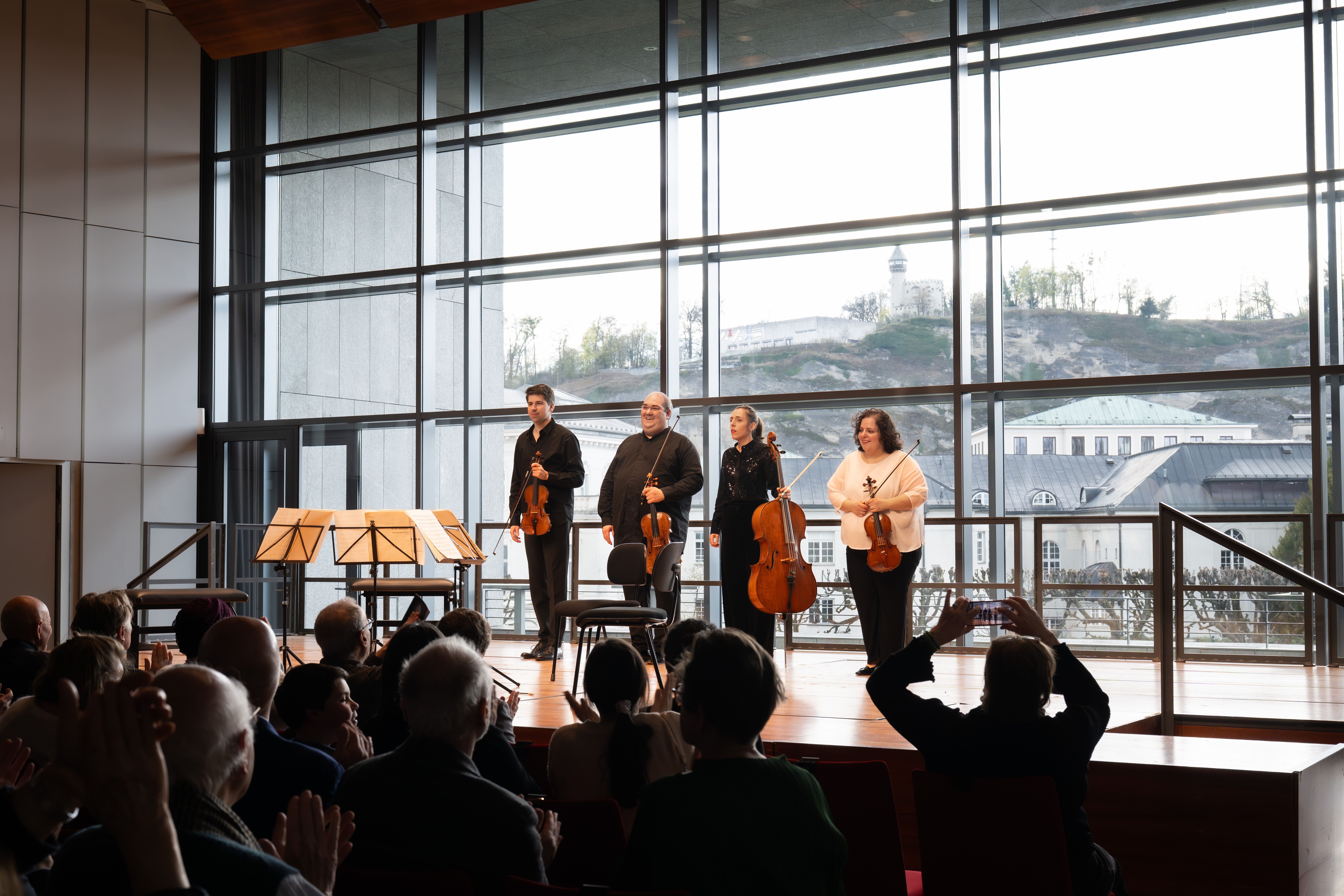 Vier Musiker mit Streichinstrumenten stehen auf der Bühne und blicken auf ein applaudierendes Publikum in einem modernen Konzertsaal mit großen Fenstern und einer malerischen Aussicht auf Gebäude und Grünflächen.