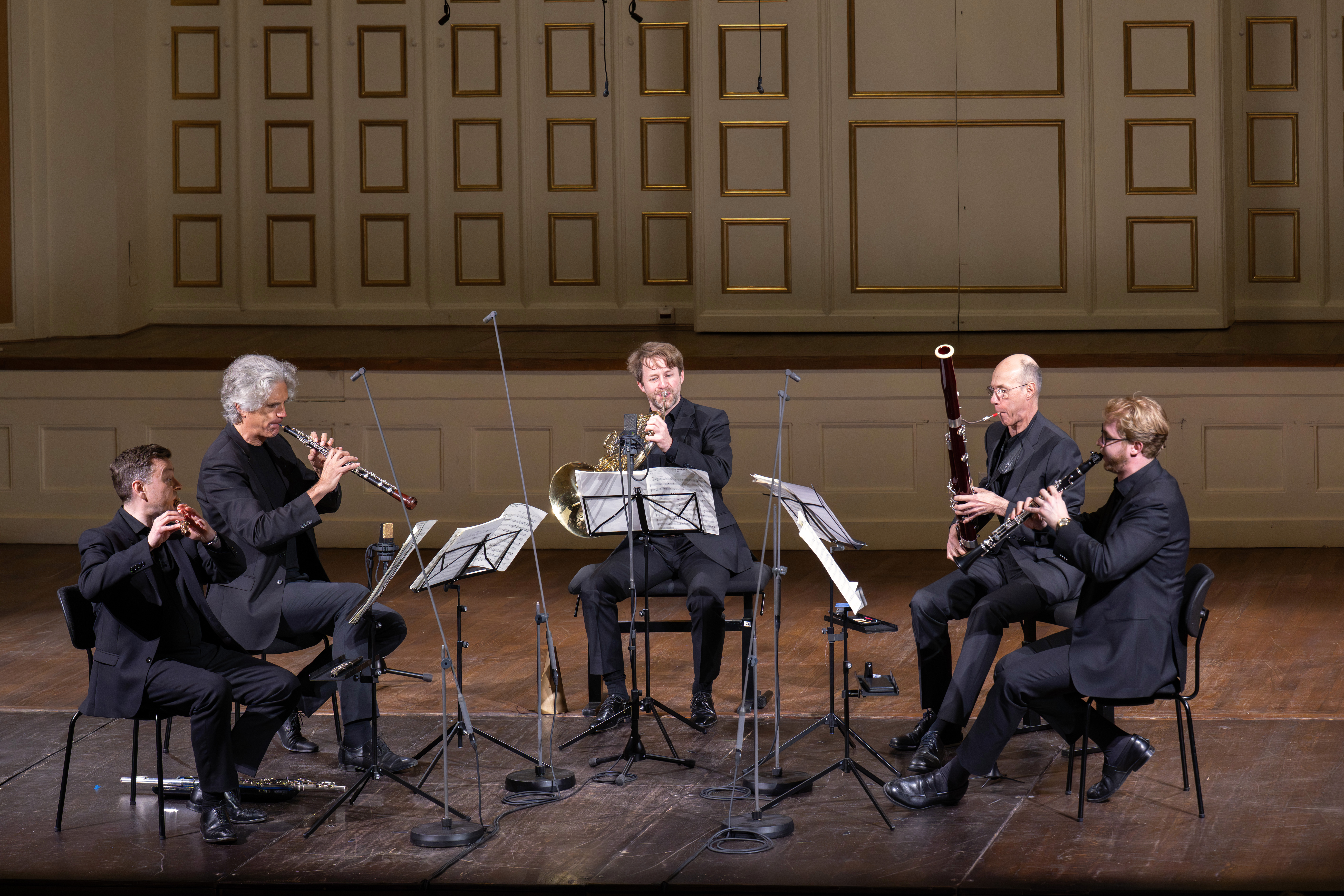 Fünf Musiker in schwarzen Anzügen treten auf einer hölzernen Bühne auf, jeder spielt ein Blasinstrument und sitzt in einem Halbkreis mit Notenständern und Mikrofonen vor ihnen, vor einem dekorierten Wandhintergrund.