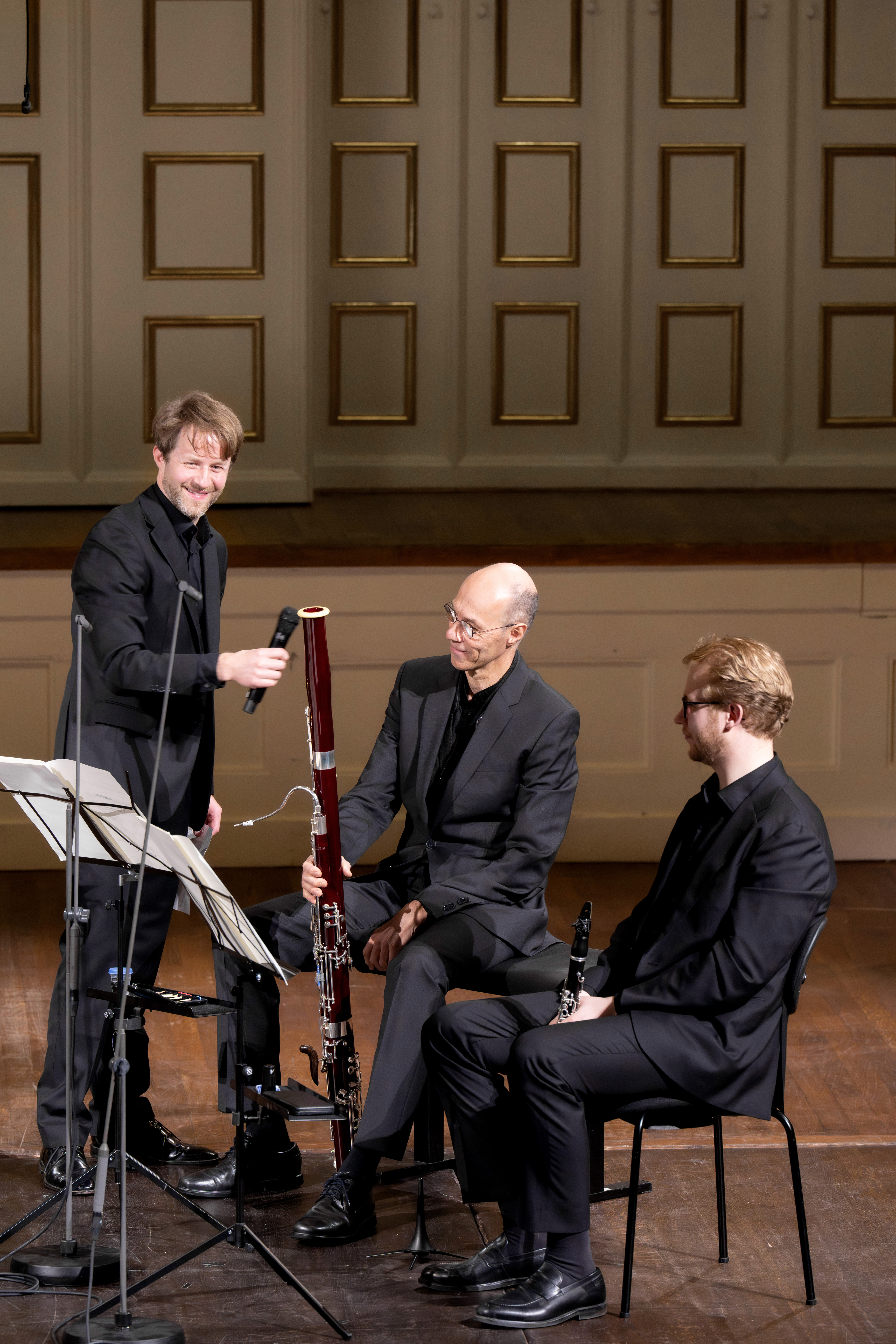 Drei männliche Musiker in schwarzen Anzügen sitzen und stehen auf einer Bühne mit Holzverkleidung. Einer hält ein Fagott, während die beiden sitzenden Männer Klarinetten halten und sich auf ihren Auftritt vorbereiten. Notenblätter liegen auf Ständern vor ihnen.