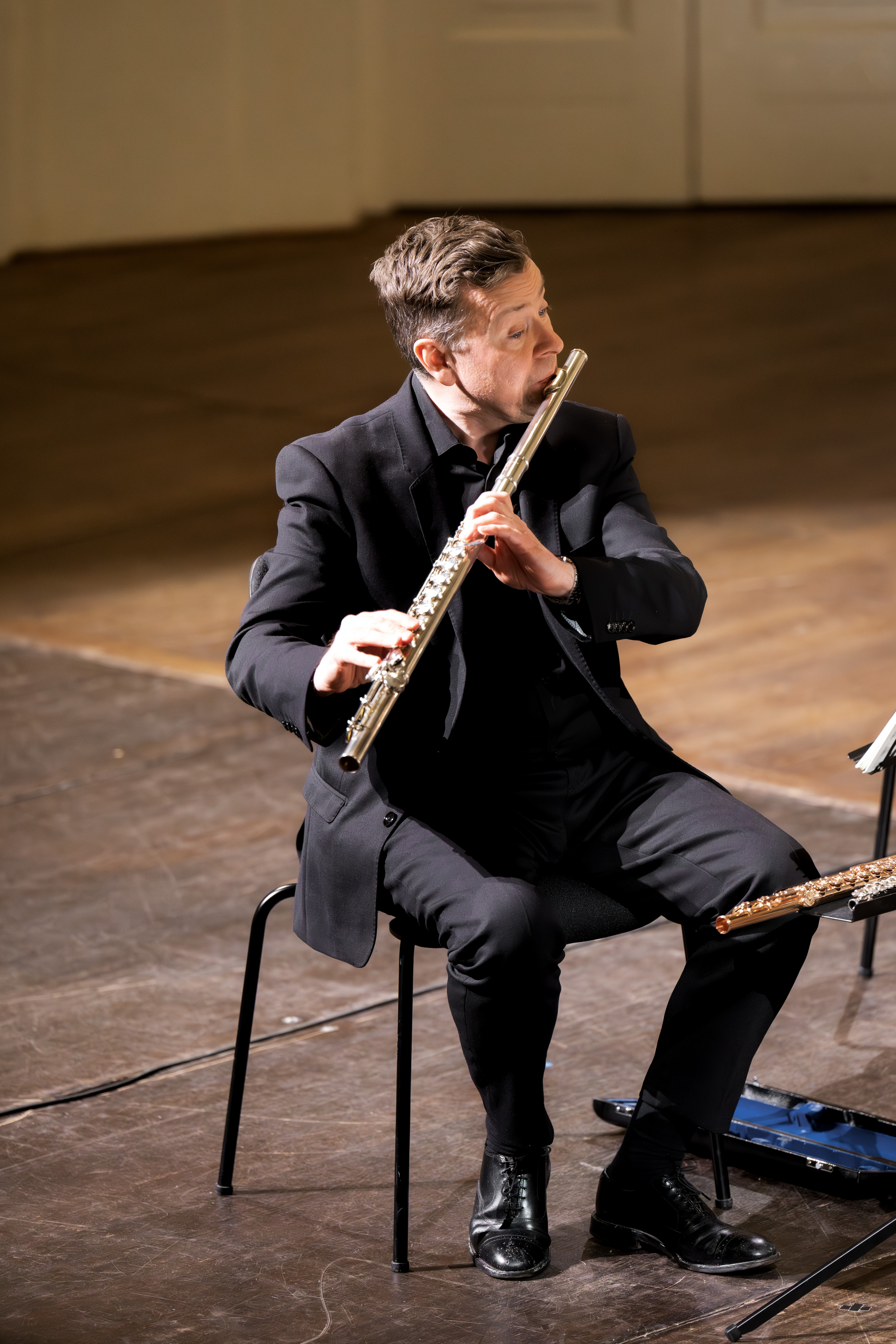 Ein Musiker im schwarzen Anzug sitzt auf einem Stuhl und spielt Flöte auf einer hölzernen Bühne. Er konzentriert sich auf seine Darbietung während eines scheinbar formellen Konzerts oder Liederabends.