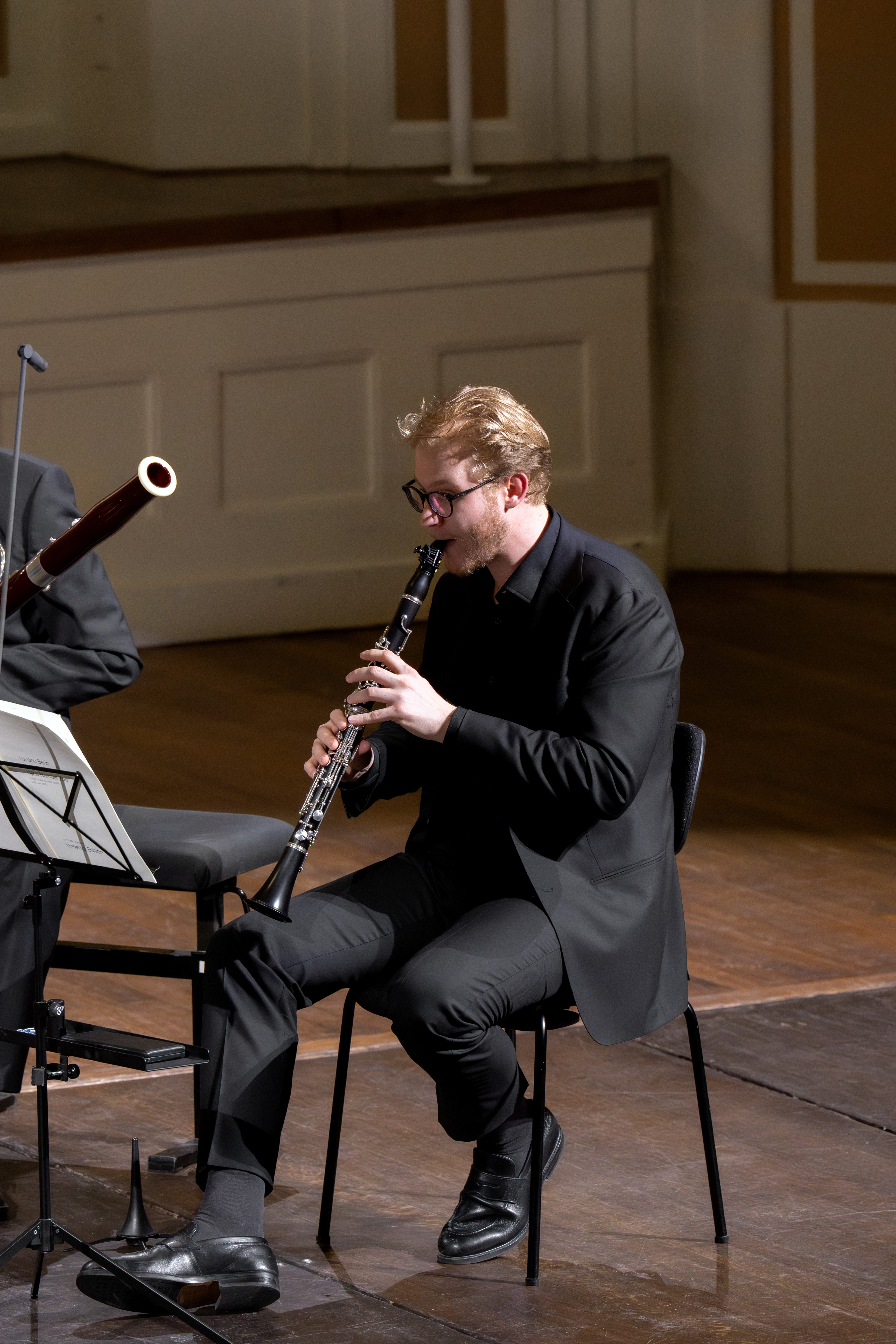 Ein Mann in schwarzer Konzertkleidung sitzt auf einem Stuhl und spielt auf der Bühne eines Konzertsaals eine Klarinette. Neben ihm sind Notenblätter zu sehen, und links ist ein Teil eines Fagotts zu erkennen.