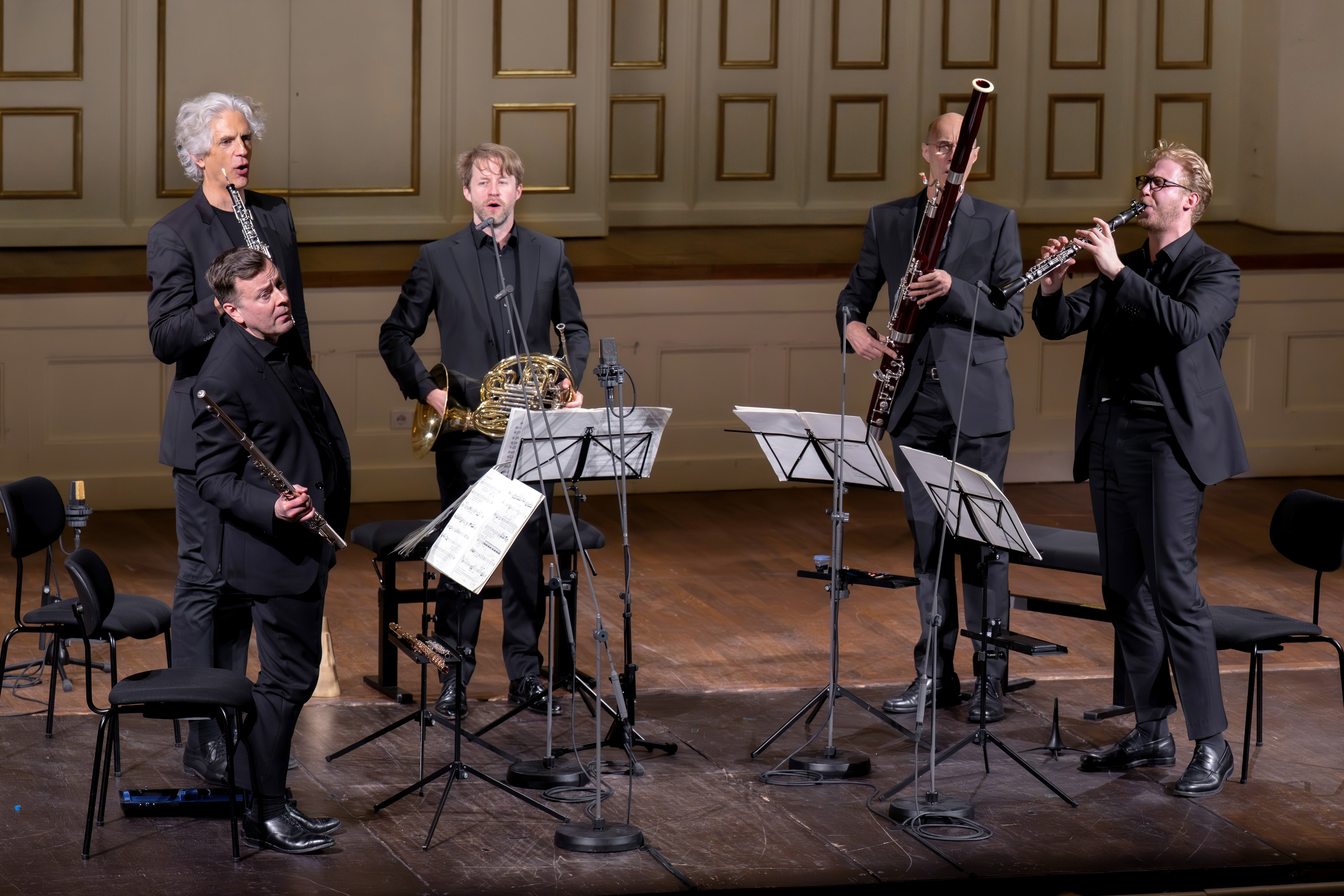 Fünf schwarz gekleidete Musiker treten auf einer Holzbühne mit Notenständern auf und spielen Blasinstrumente. Im Hintergrund ist eine dekorative Wand mit goldenen Akzenten zu sehen.