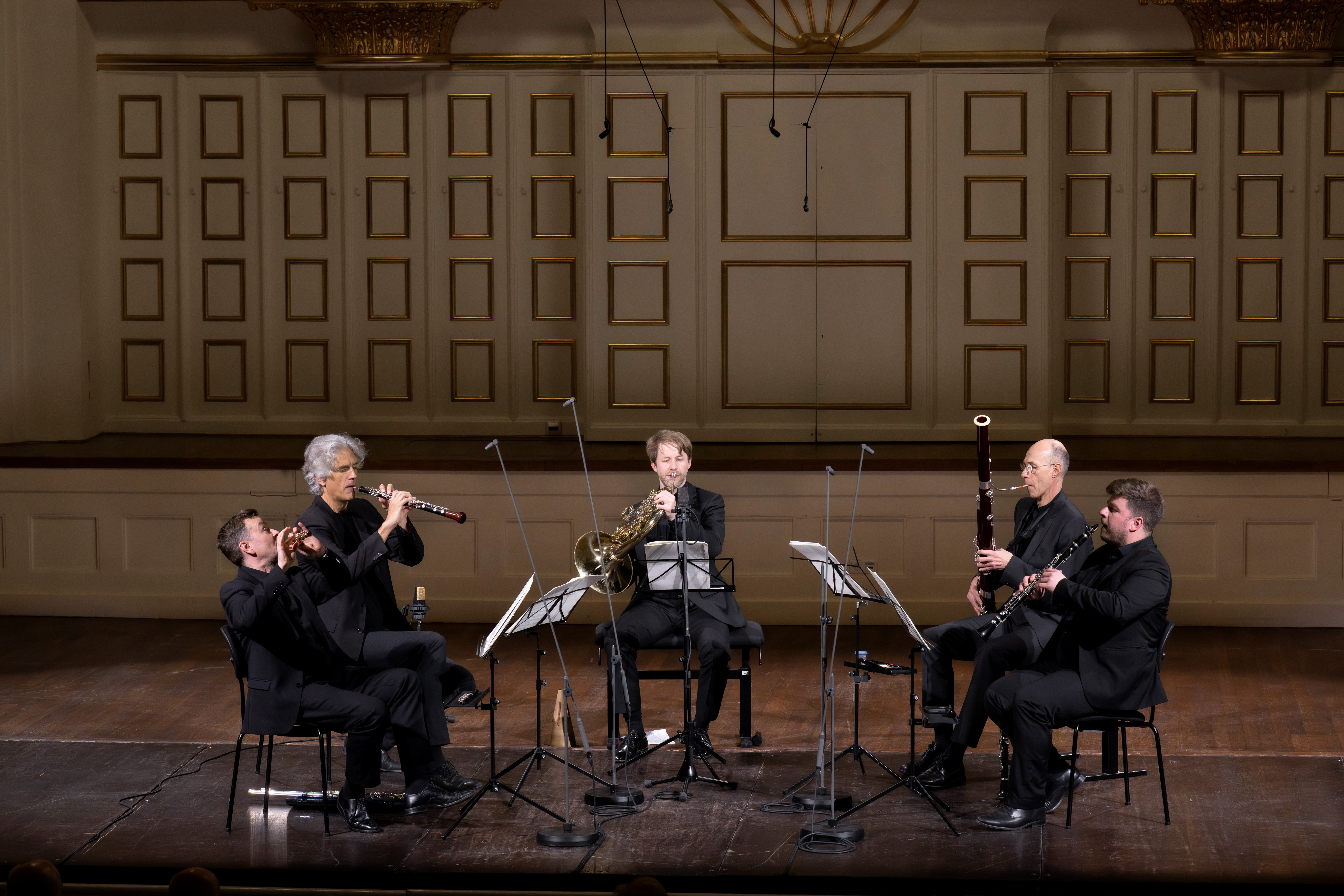 Fünf Musiker in schwarzen Anzügen spielen auf der Bühne mit Holz- und Blechblasinstrumenten, sitzen im Halbkreis und haben Notenständer und Mikrofone vor sich stehen. Verzierte Wände und Beleuchtung schmücken den Hintergrund.