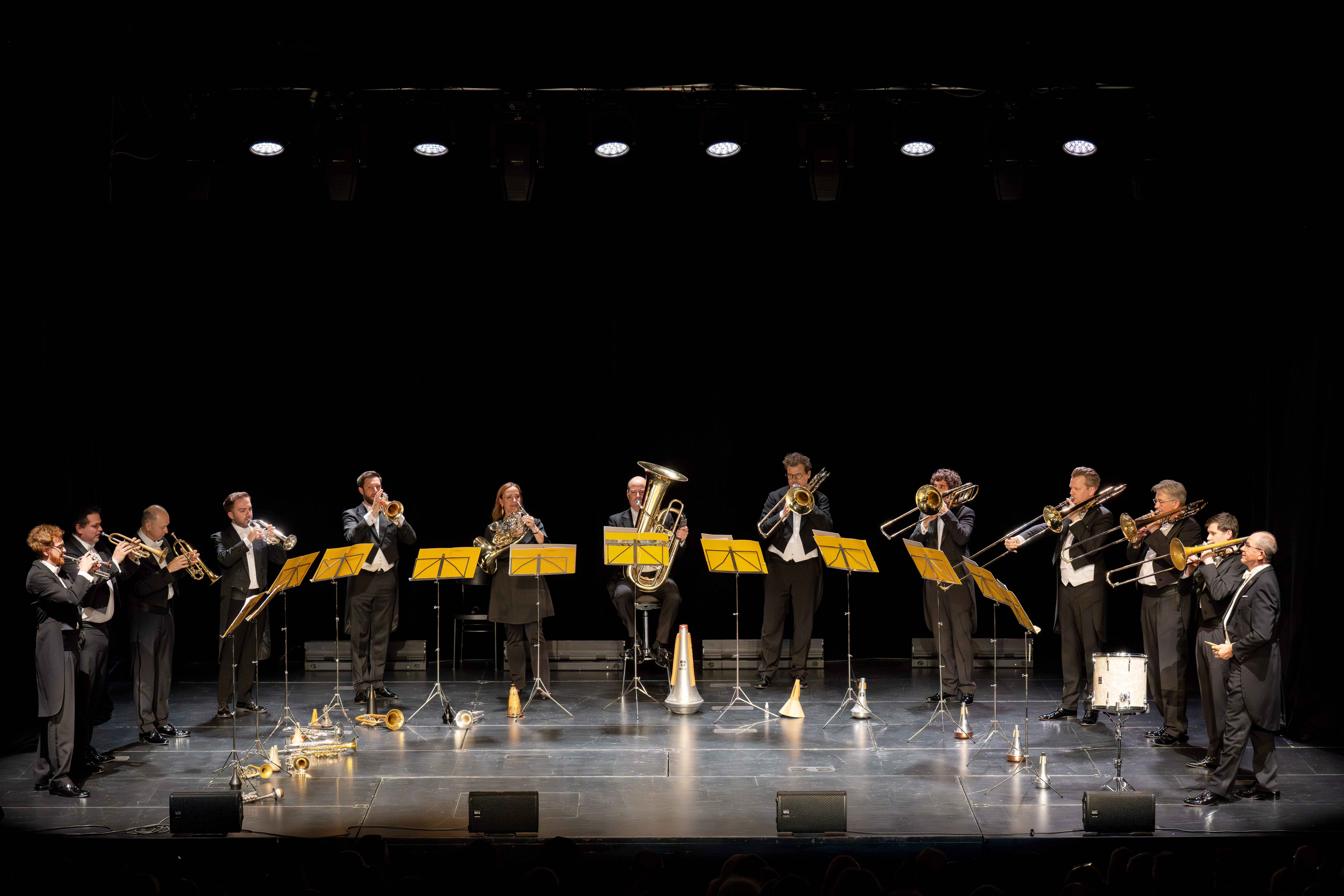 Zwölf Musiker in förmlicher Kleidung spielen auf der Bühne Blechblasinstrumente, die in einem Halbkreis hinter gelben Notenständern stehen, unter Bühnenbeleuchtung mit schwarzem Hintergrund.
