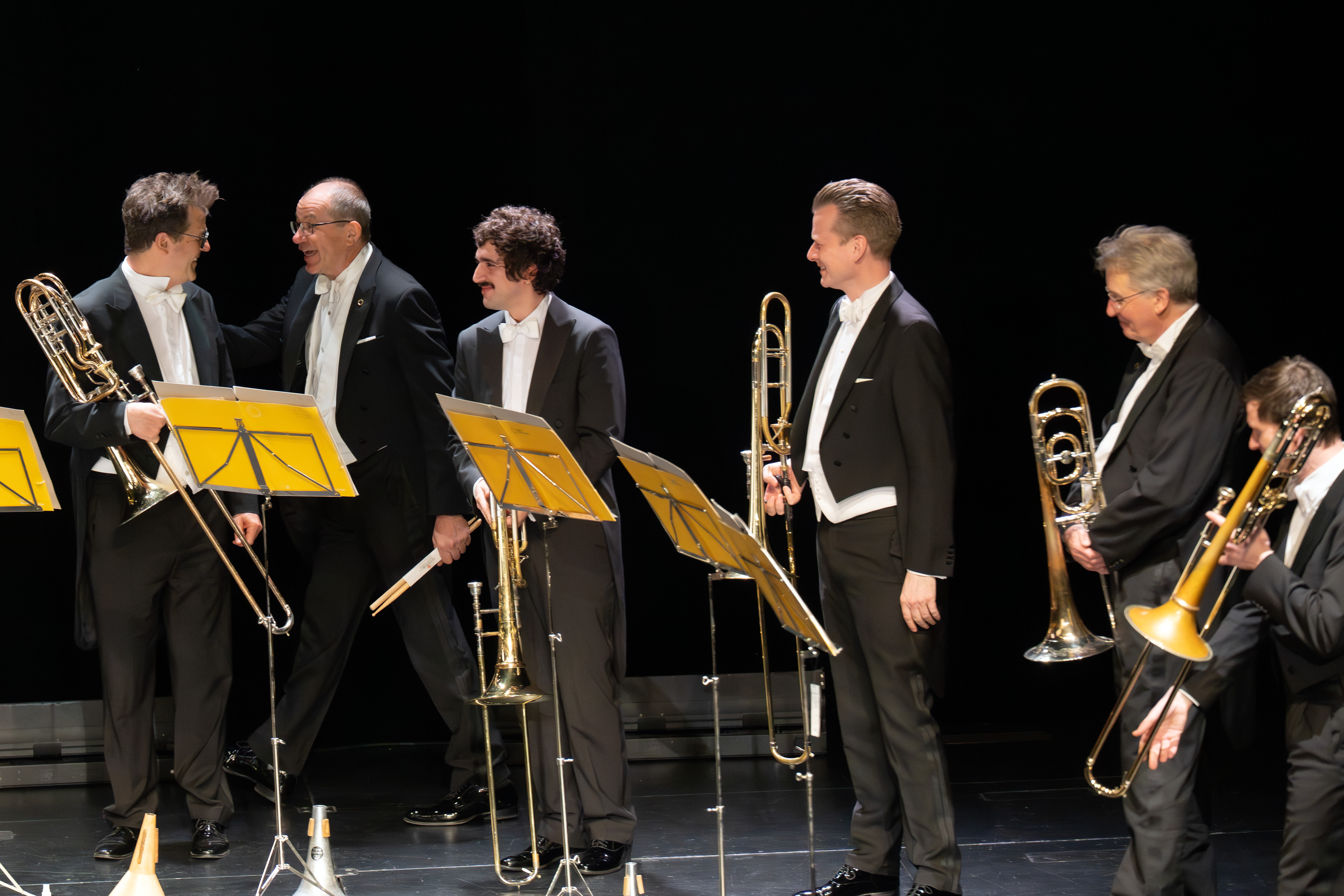 Fünf Männer in formeller schwarzer Kleidung mit Posaunen stehen auf der Bühne, lächeln und unterhalten sich, vor ihnen gelbe Notenständer, dahinter ein schwarzer Hintergrund.