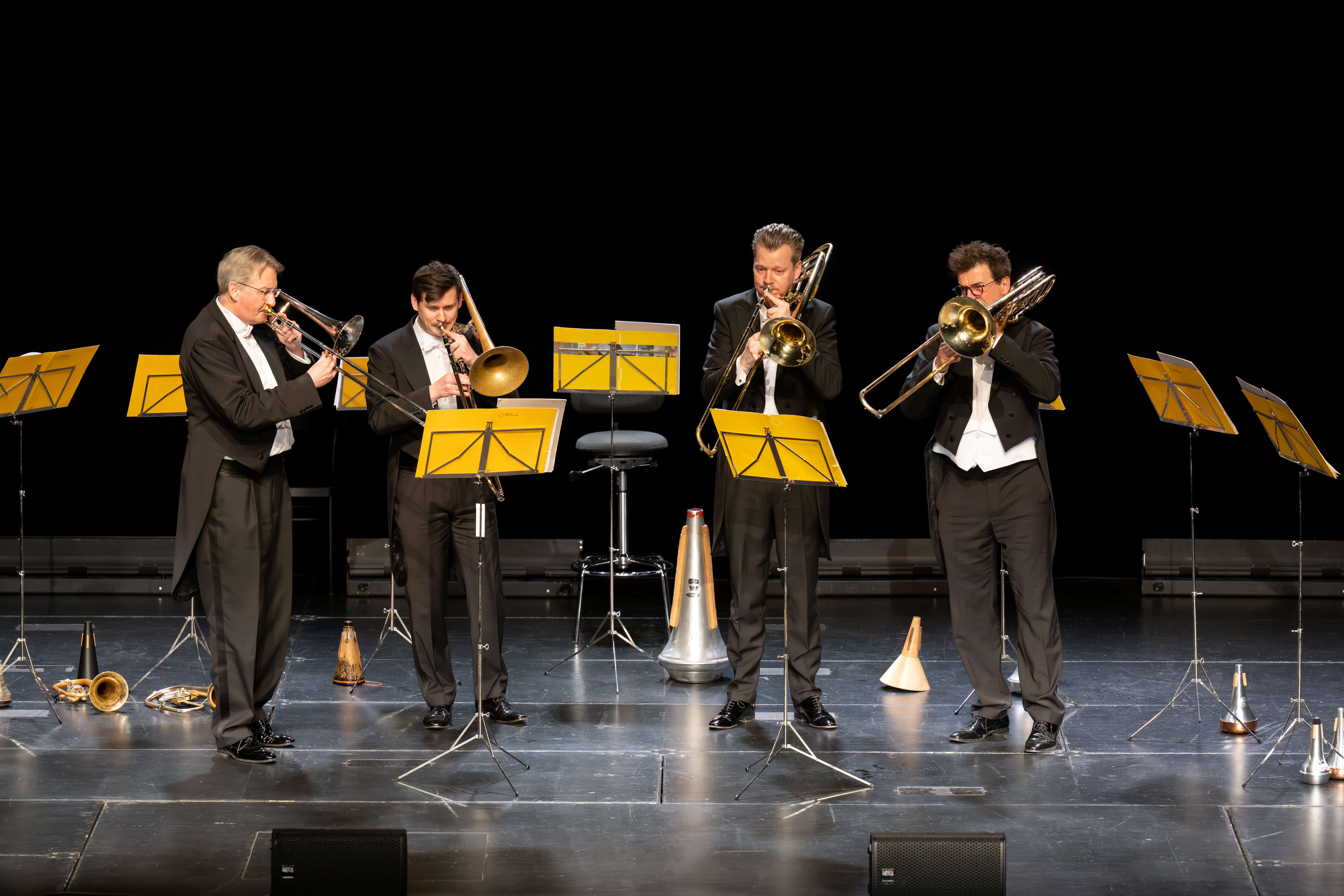 Vier Musiker in Smokings spielen auf der Bühne Posaunen, jeder steht hinter einem gelben Notenpult. Um sie herum liegen verschiedene Blechblasinstrumente und Dämpfer auf dem Boden. Schwarzer Hintergrund.