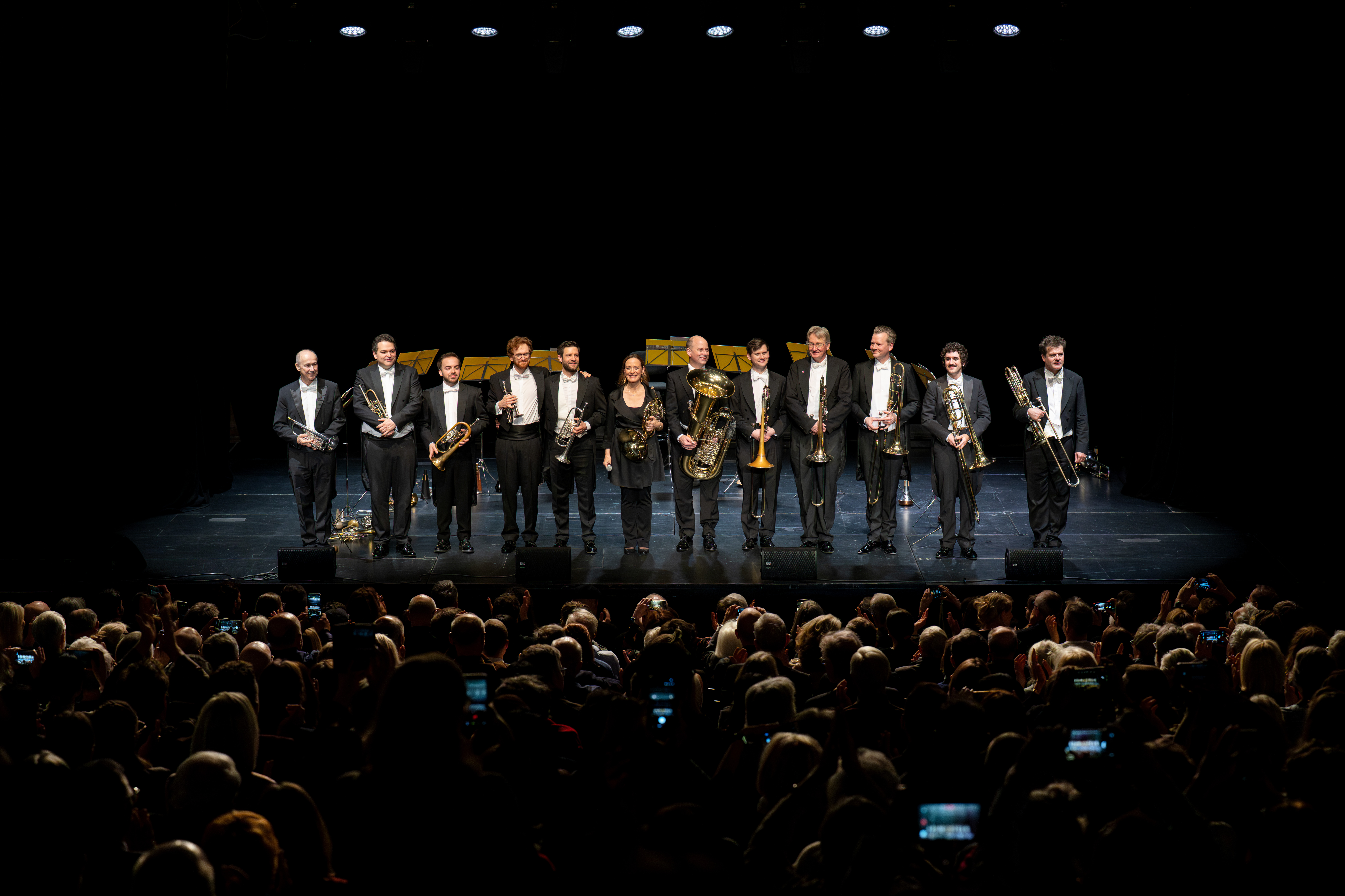 Eine Gruppe von Musikern in förmlicher Kleidung steht auf einer Bühne mit Blechblasinstrumenten und verbeugt sich vor dem Publikum in einem dunklen Theater, während die Zuschauer von ihren Plätzen aus Fotos machen.
