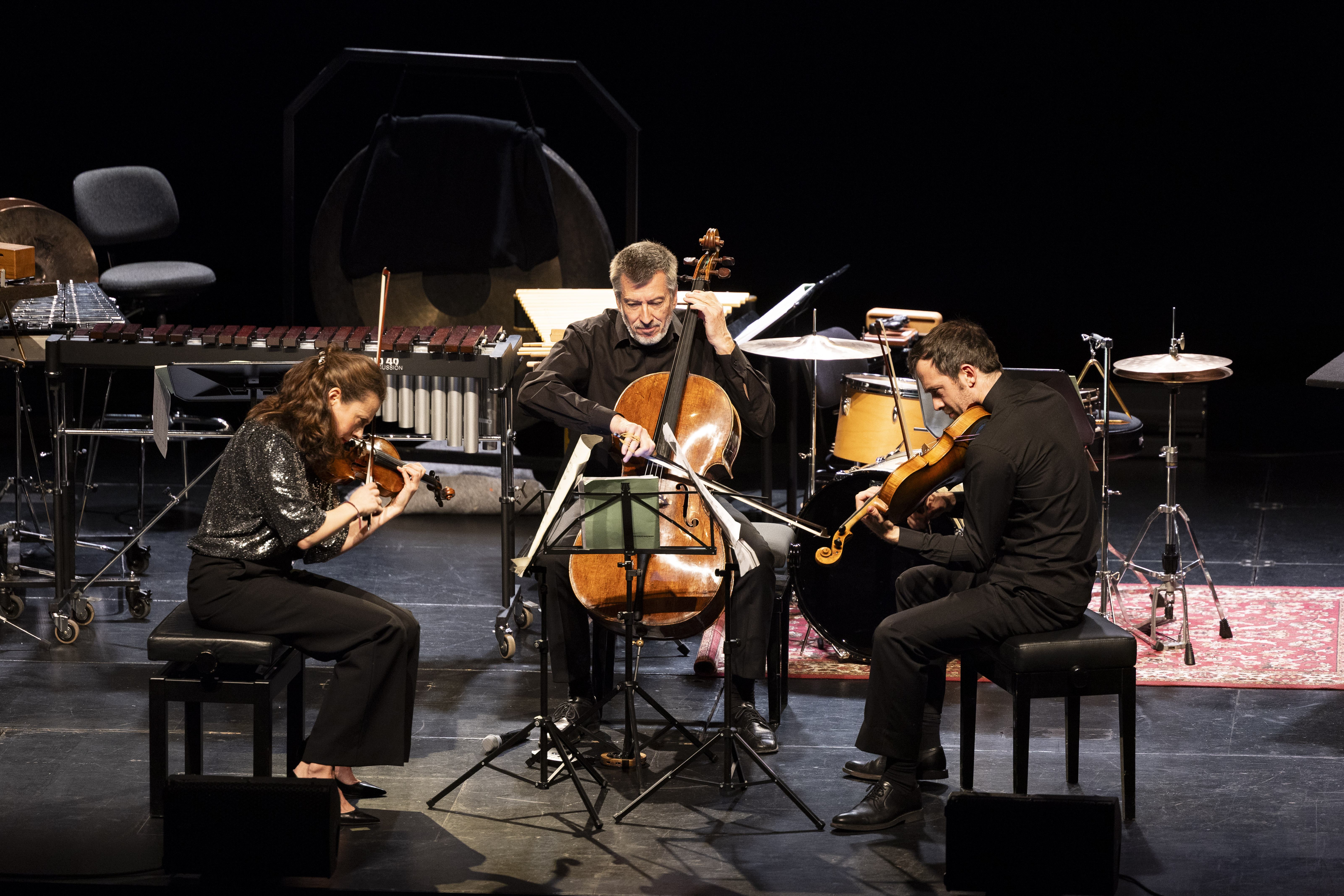 Drei Musiker spielen auf der Bühne: eine Frau, die Geige spielt, ein Mann, der Cello spielt, und ein weiterer Mann, der Bratsche spielt. Im Hintergrund sind Perkussionsinstrumente und Notenständer zu sehen.