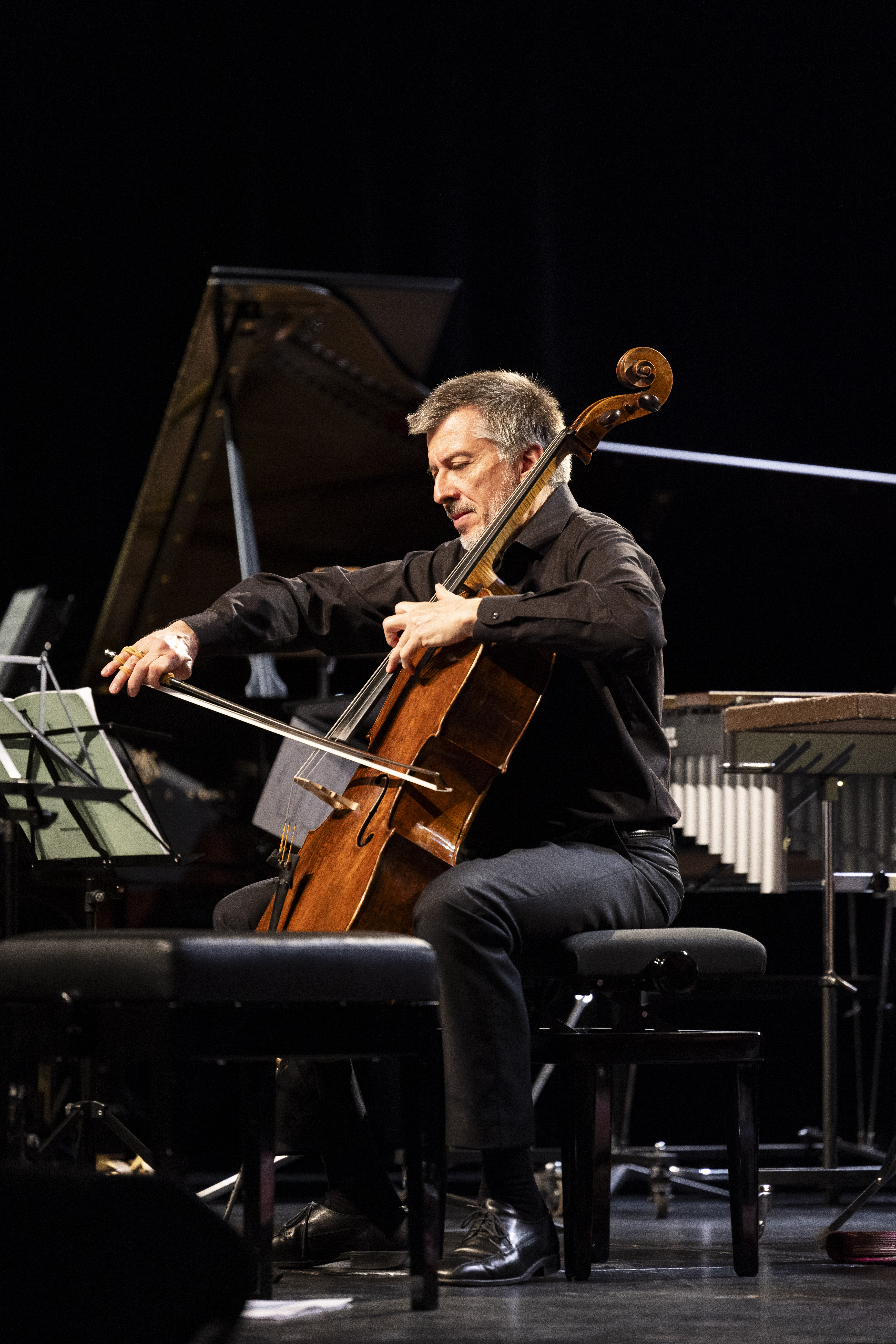Ein Mann in schwarzer Kleidung spielt auf der Bühne vor Notenblättern sitzend Cello, während im Hintergrund ein Flügel und Schlaginstrumente zu sehen sind.