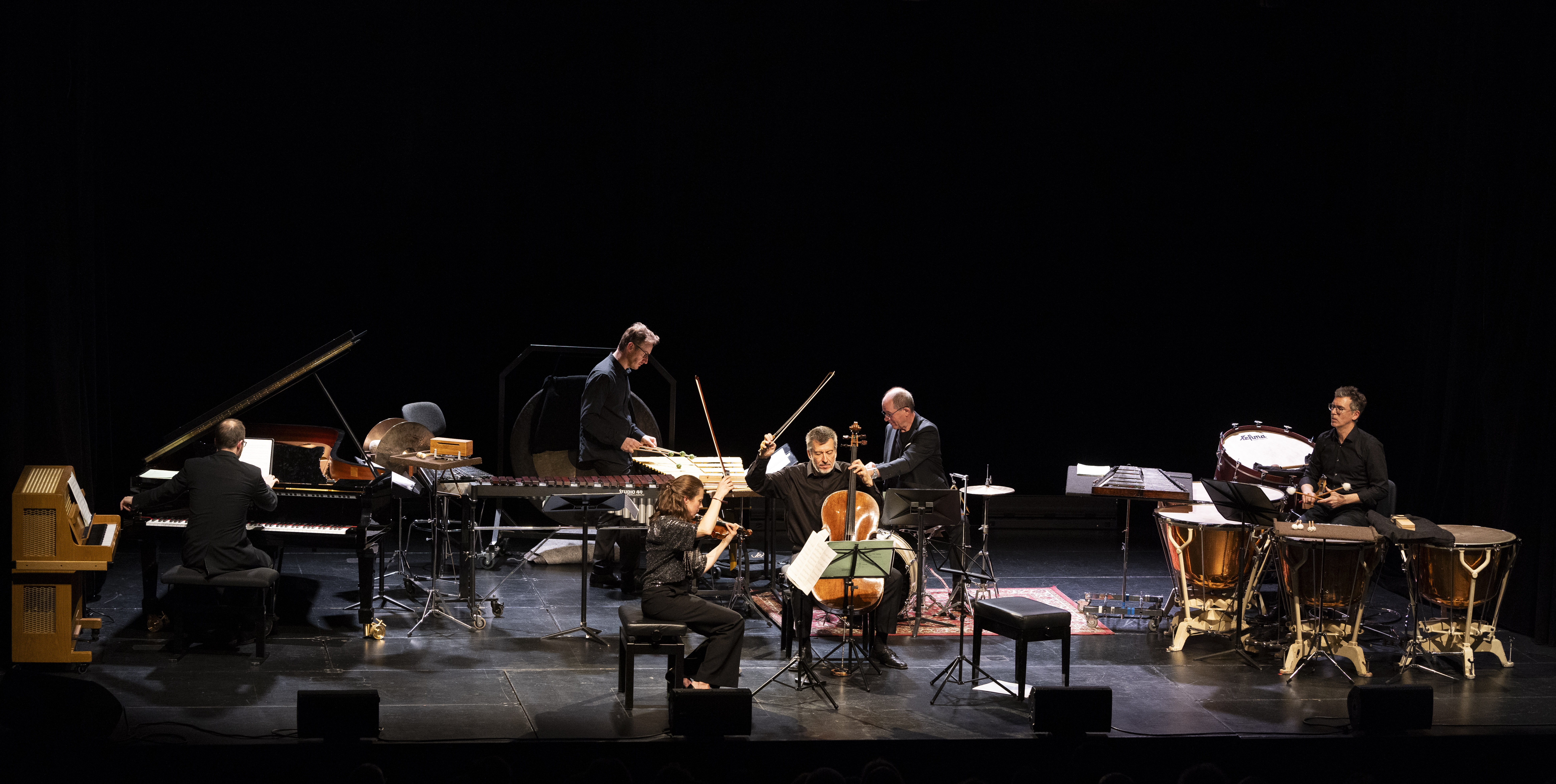 Sechs Musiker spielen auf der Bühne mit Klavier, Streichern, Schlagzeug und anderen Instrumenten. Sie sind schwarz gekleidet, sitzen im Halbkreis und sind vor einem dunklen Hintergrund beleuchtet.