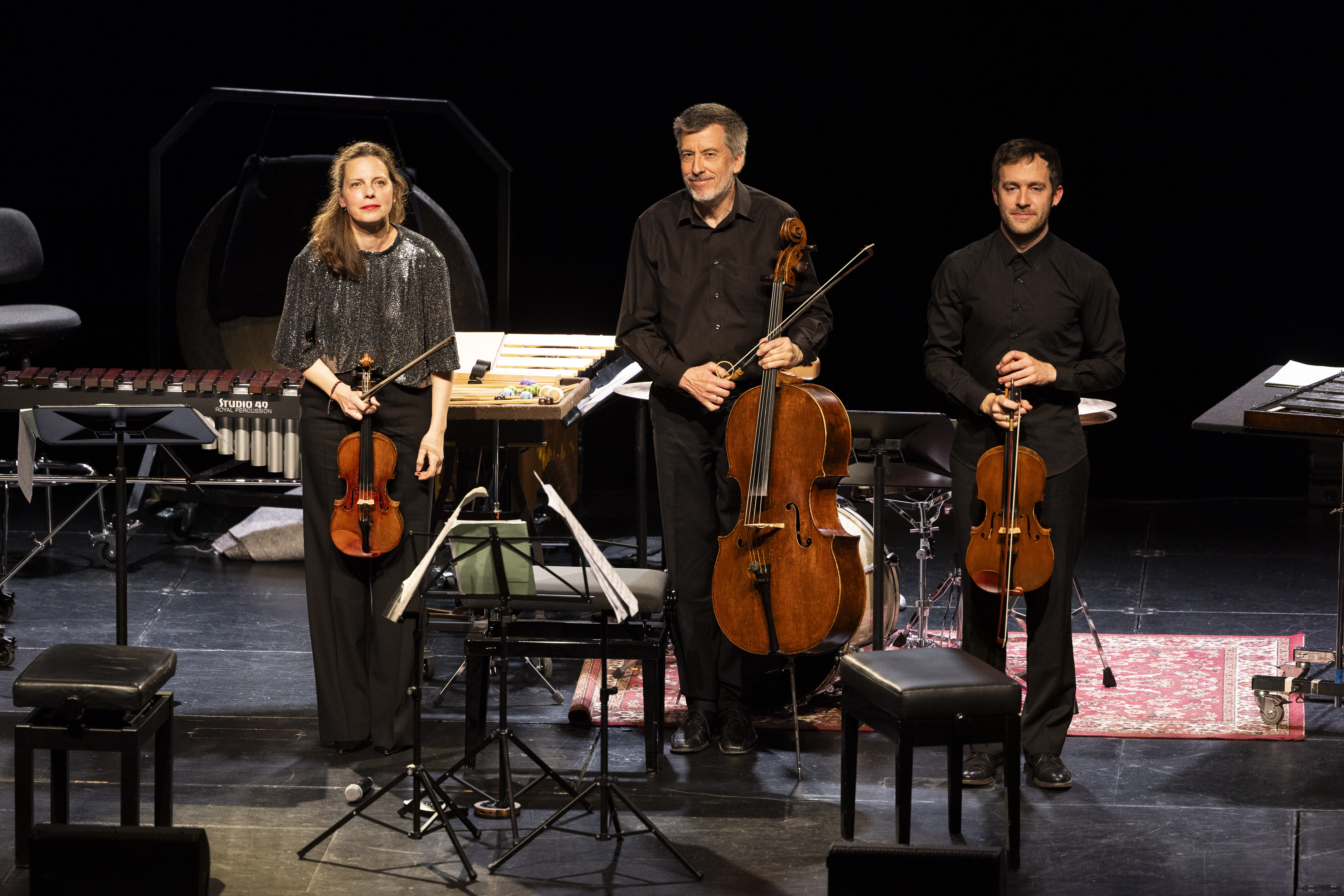 Drei Musiker stehen auf der Bühne und halten ihre Streichinstrumente, zwei Geigen und ein Cello, während im Hintergrund Notenständer, Stühle und Schlaginstrumente zu sehen sind.