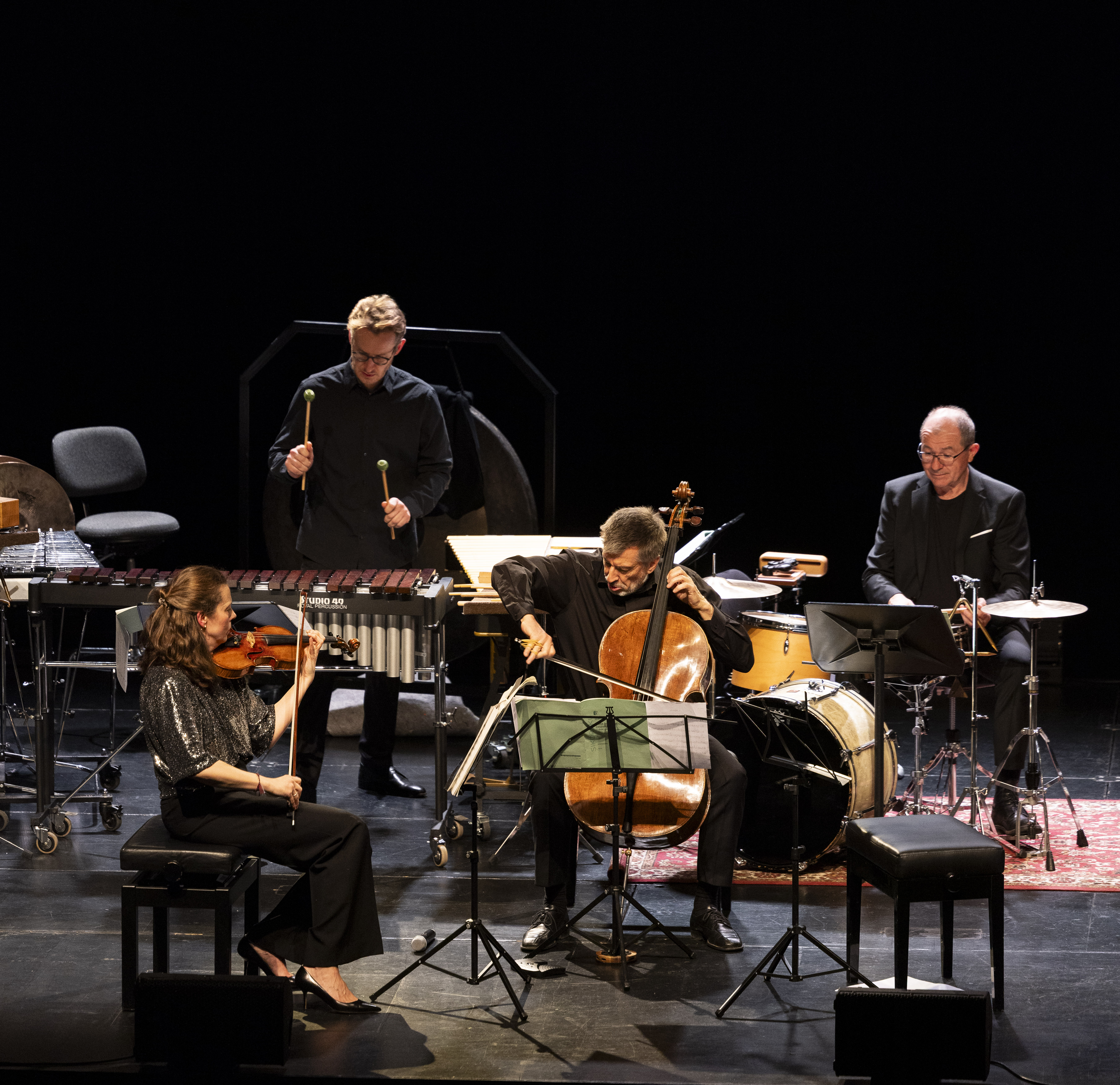 Vier Musiker treten auf der Bühne auf: Vorne sitzen ein Geiger und ein Cellist, dahinter ein Schlagzeuger mit Schlägeln und ein Schlagzeuger, umgeben von ihren Instrumenten. Der Hintergrund ist dunkel.