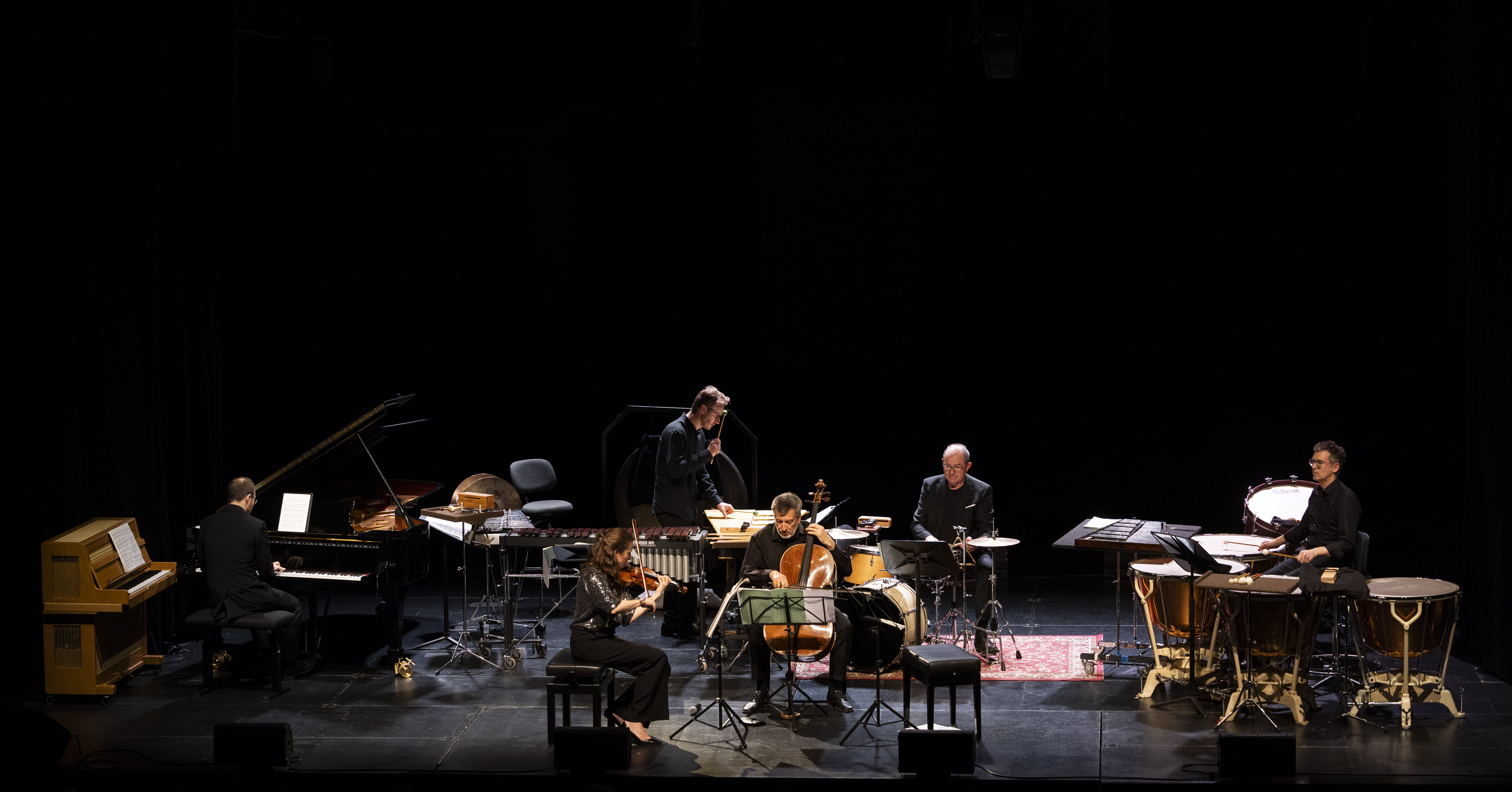 Ein kleines Orchester mit sechs Musikern, die auf der Bühne Klavier, Violine, Cello, Kontrabass, Schlagzeug und Pauken spielen, wobei die Notenständer und Instrumente in einem Halbkreis angeordnet sind.