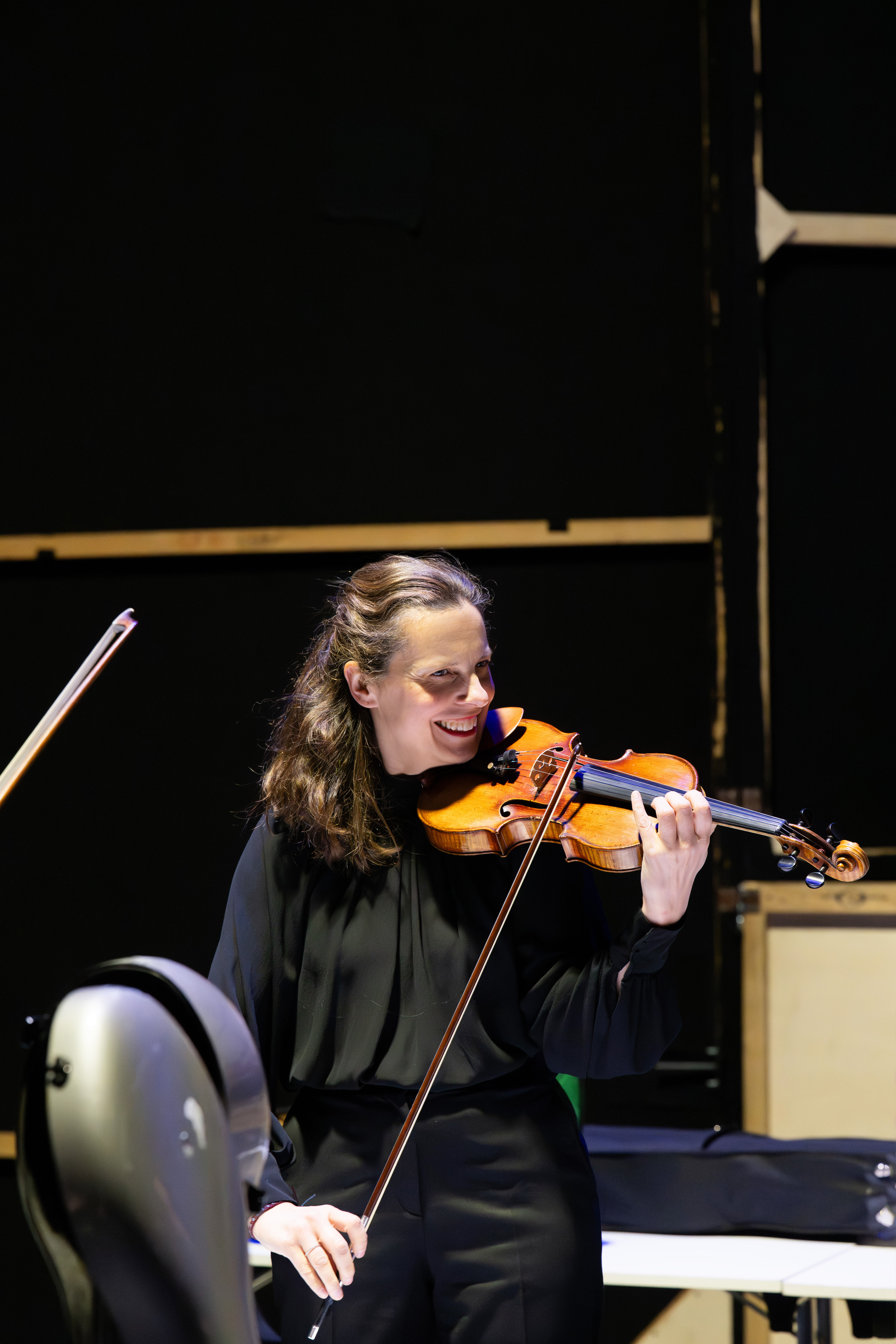 A smiling woman in a black outfit plays a violin on stage, with music stands and a cello visible nearby against a dark background.