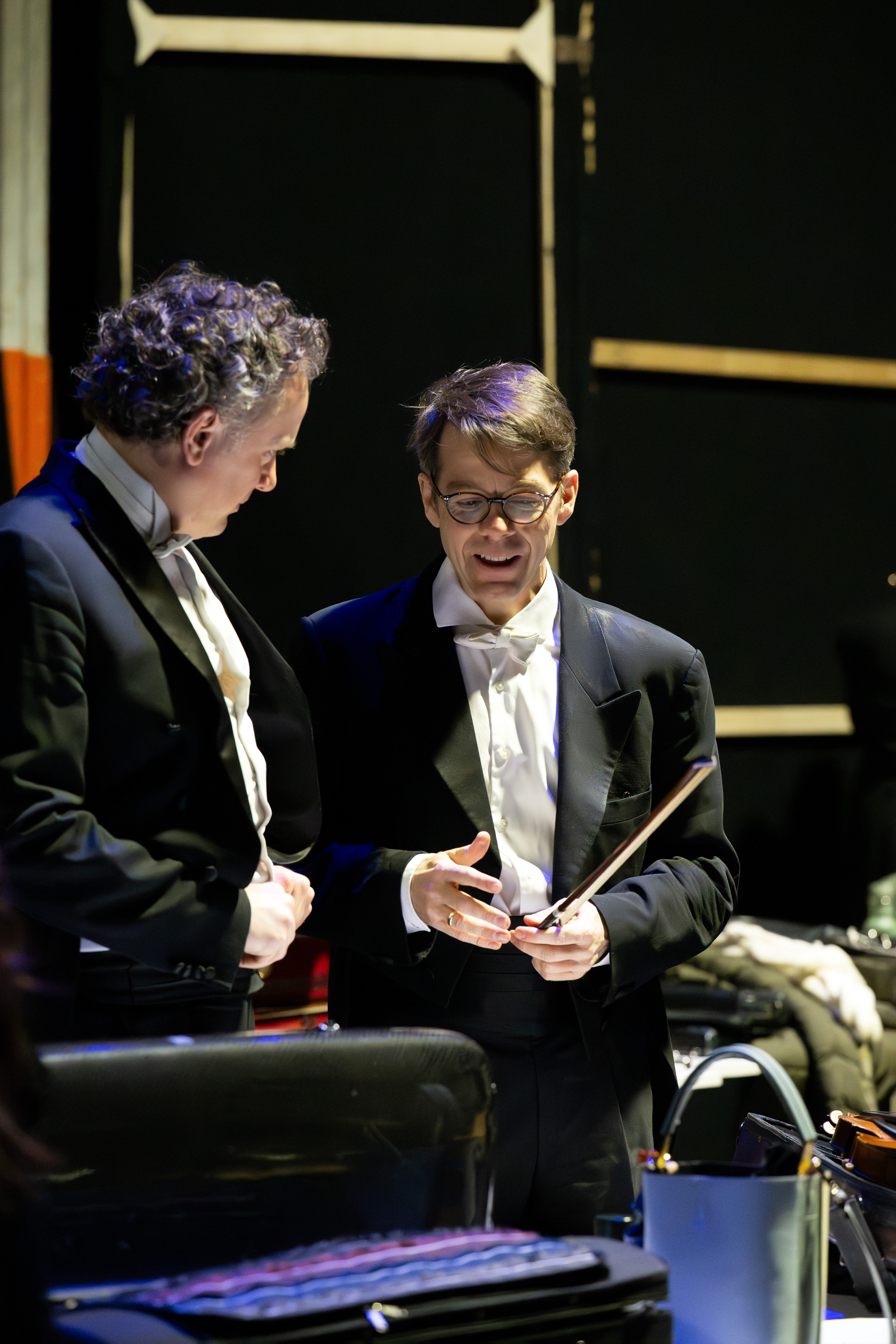 Two men in formal black tuxedos stand and converse backstage, one holding a conductor’s baton, with musical instruments and cases visible nearby.
