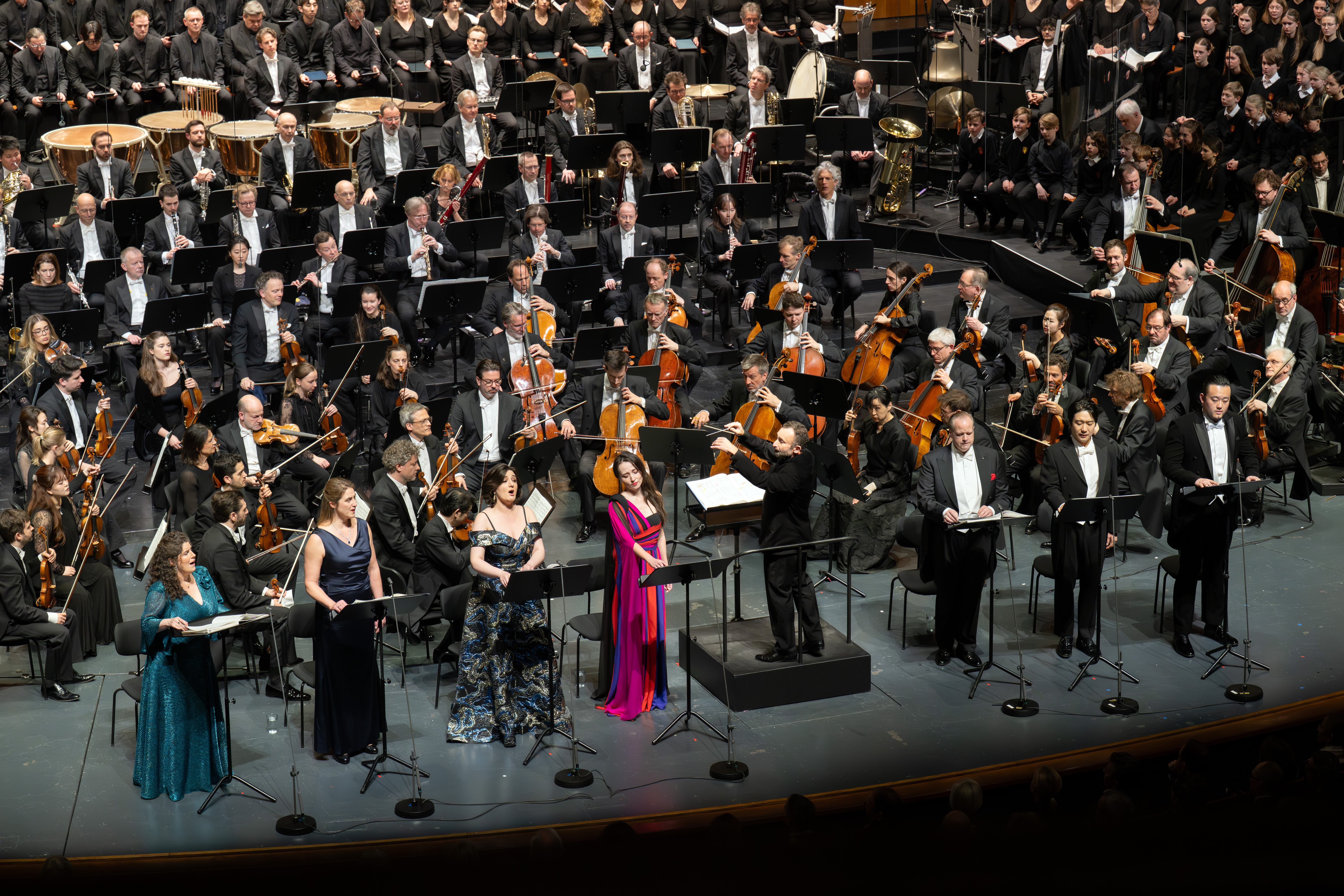Ein großes Orchester und ein Chor treten auf der Bühne auf. Sechs Solisten stehen vorne neben einem Dirigenten, alle formell gekleidet. Hinter ihnen sitzen die Musiker mit Streich- und Blasinstrumenten.