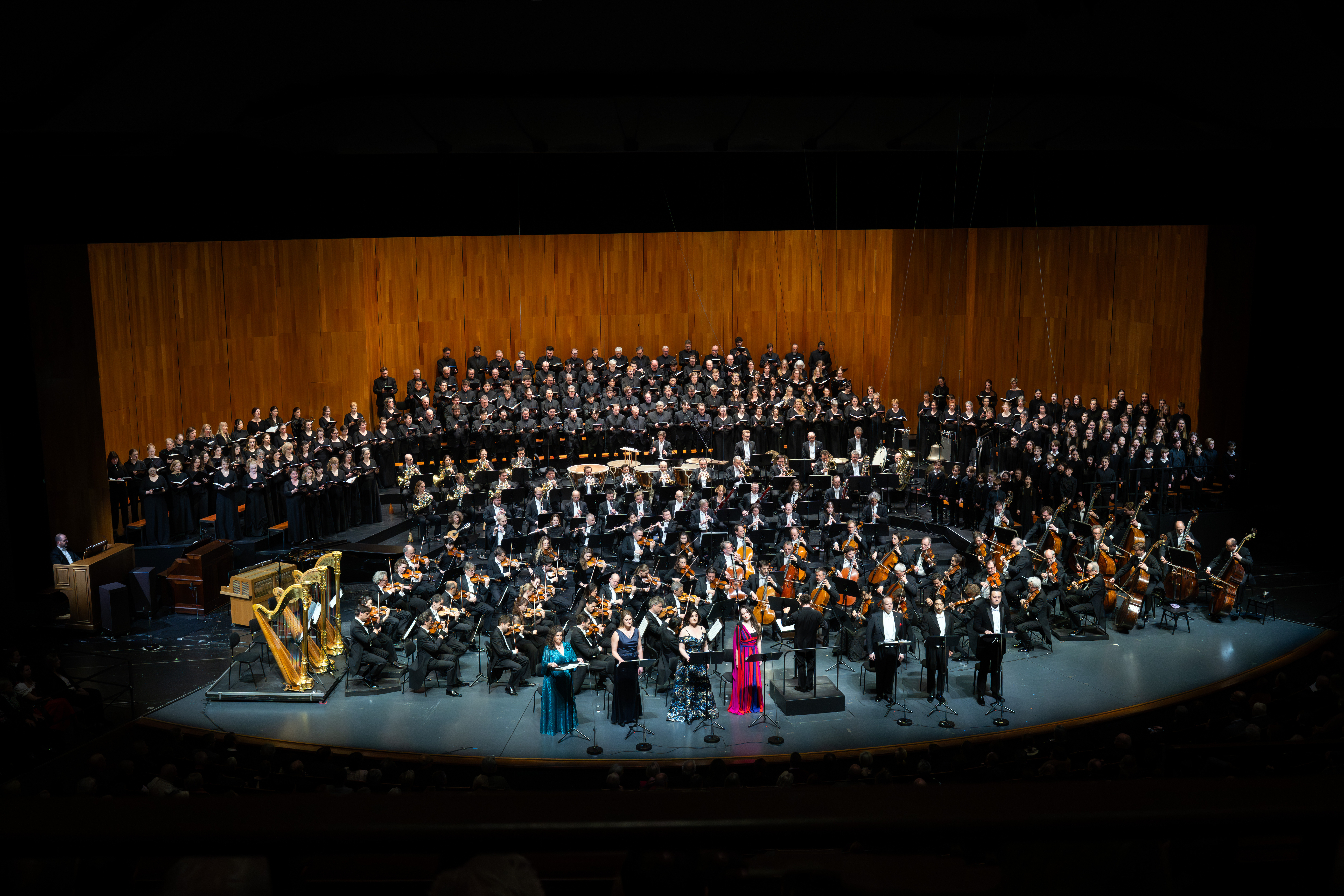 Ein großes Orchester und ein Chor treten auf einer gut beleuchteten, holzgetäfelten Bühne auf. Mehrere Solisten stehen vorne, einer in einem leuchtend rosafarbenen Kleid, während das Publikum aus dem abgedunkelten Theater zuschaut.
