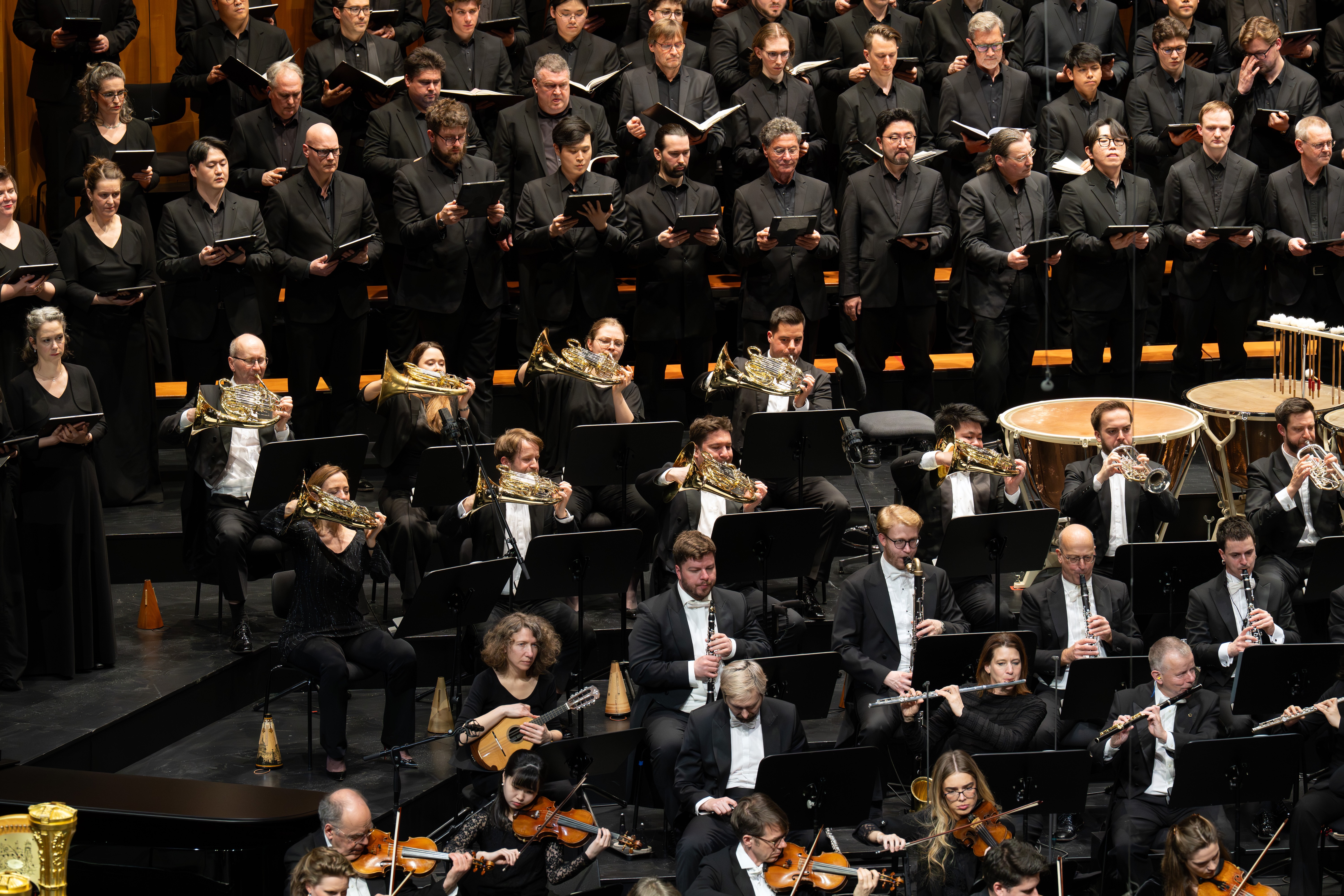 Ein großes Orchester und ein Chor treten auf der Bühne auf. Die Musiker spielen verschiedene Instrumente, und der Chor steht in Reihen hinter ihnen, alle in formellem schwarzen Gewand.