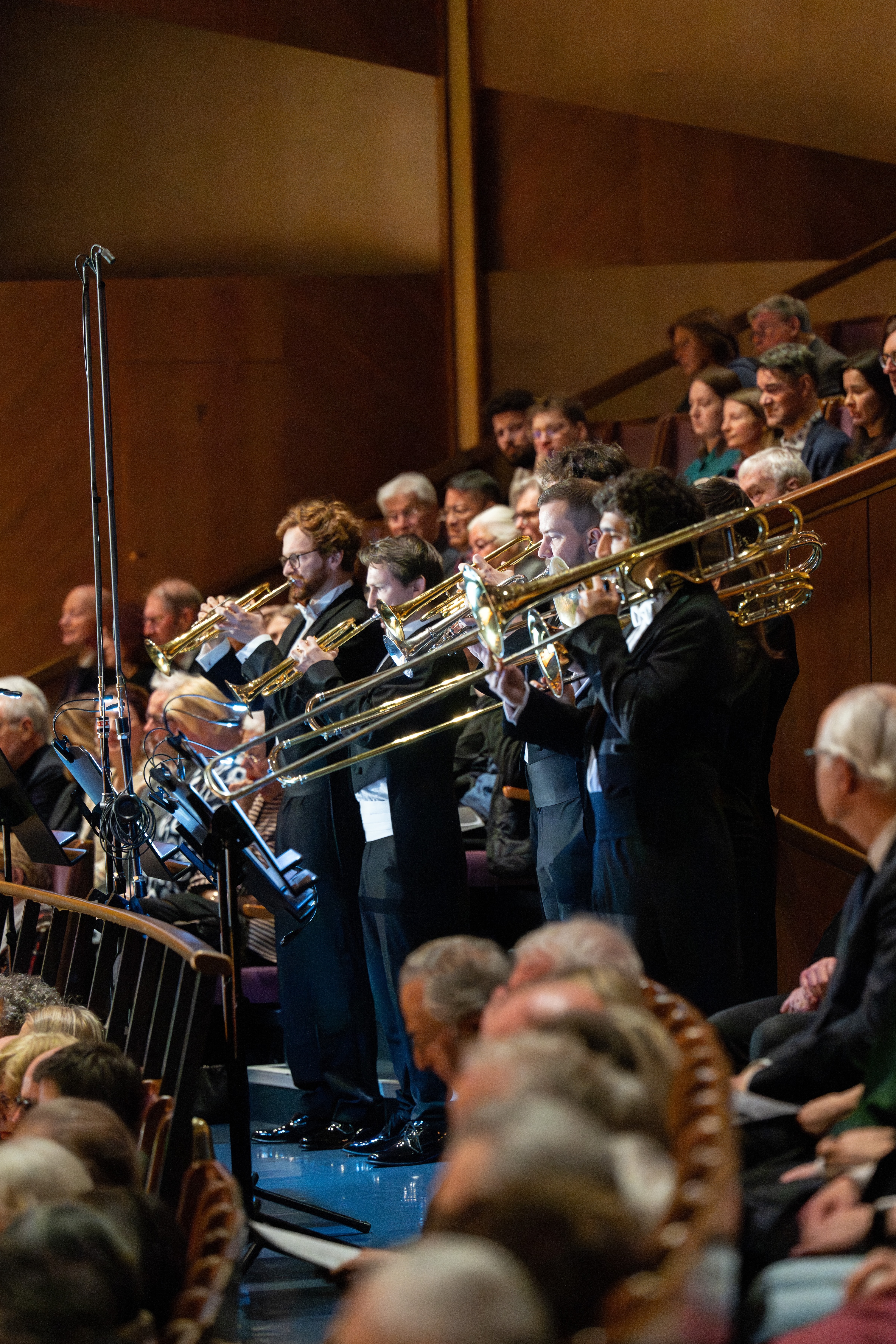 Trombone players in formal attire perform in an orchestra, standing among seated musicians and an audience in a concert hall with wooden walls and tiered seating.