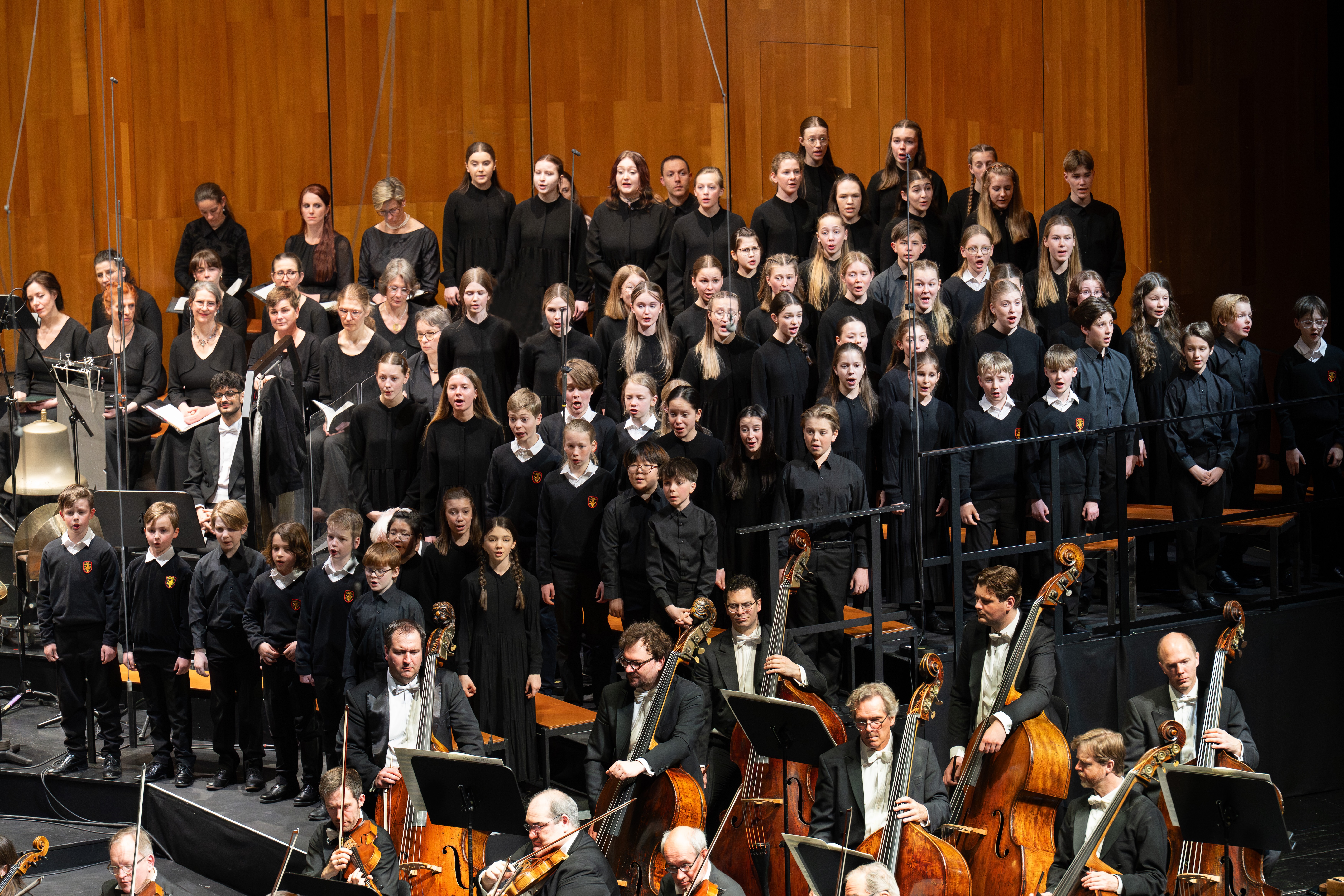 Ein großer Chor von Kindern in schwarzer Kleidung steht auf Steigbügeln hinter einem Orchester aus sitzenden Musikern, die auf einer hölzernen Konzertsaalbühne Streichinstrumente spielen.