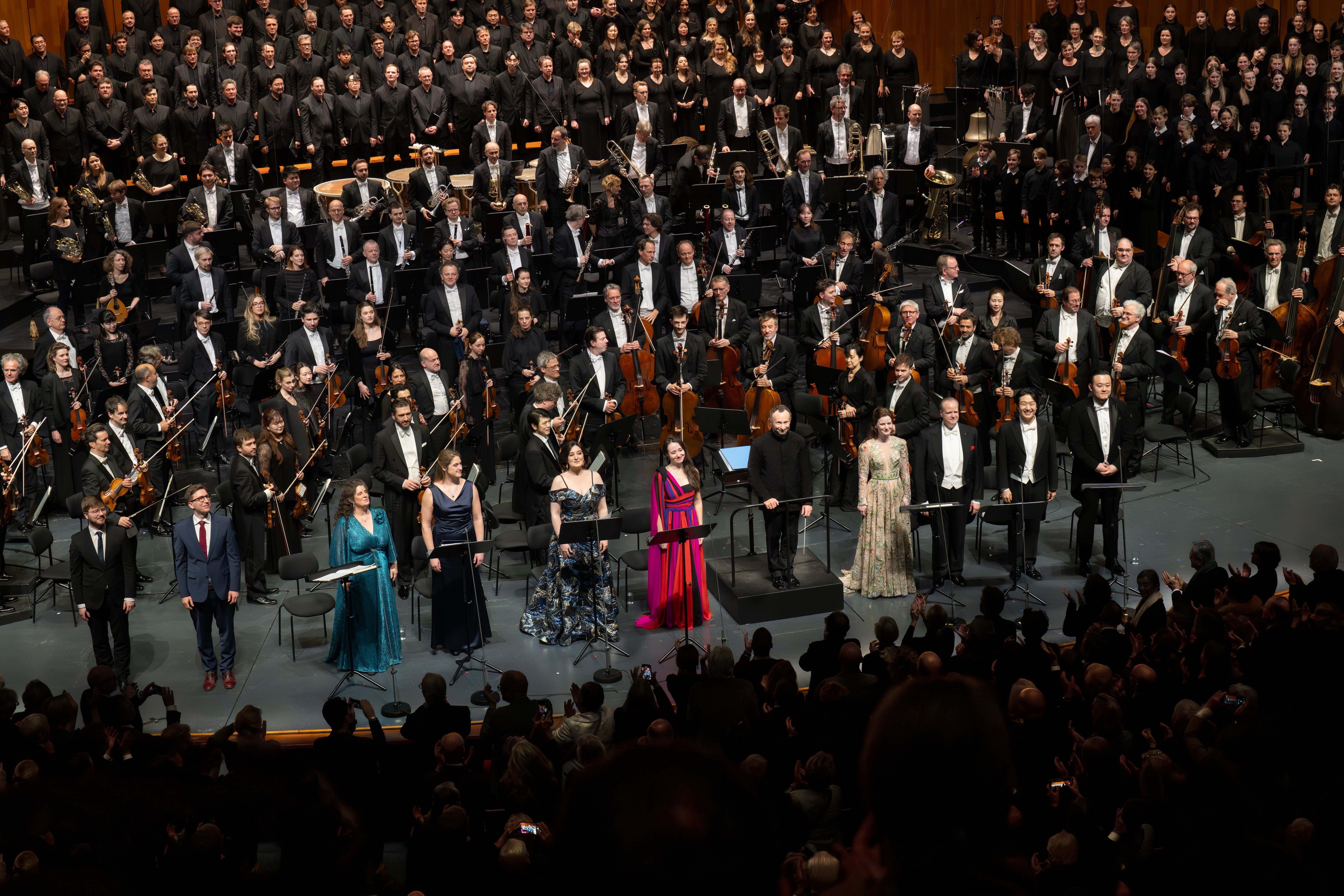Auf der Bühne treten ein großes Orchester und ein Chor auf, mit mehreren Solisten und einem Dirigenten, die alle formell gekleidet sind; im Vordergrund ist das Publikum zu sehen.