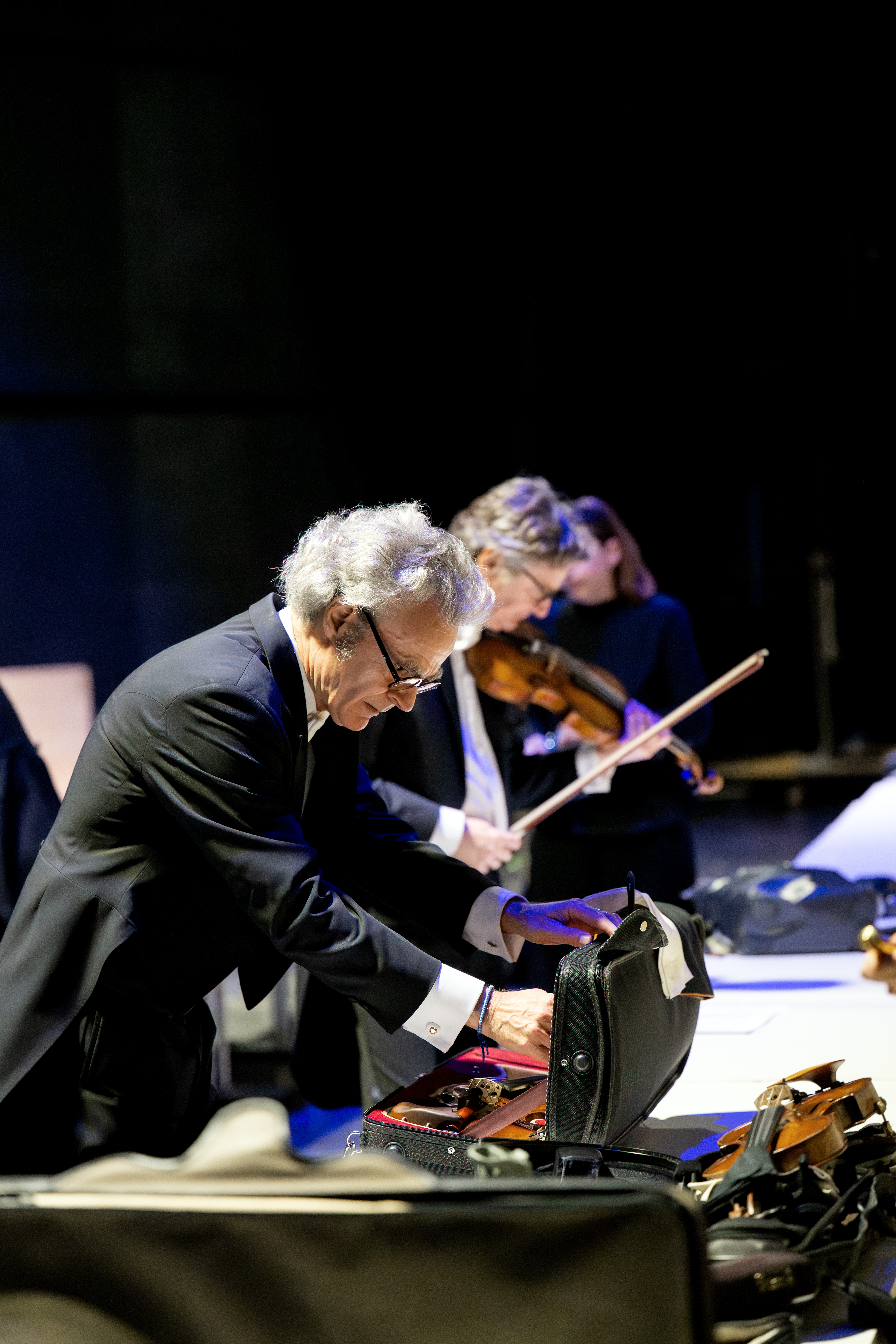 Ein Mann im Anzug öffnet einen Geigenkasten auf einem Tisch, während eine Frau im Hintergrund eine Geige spielt; beide befinden sich in einer schwach beleuchteten, formellen Umgebung, möglicherweise hinter der Bühne eines Konzerts.