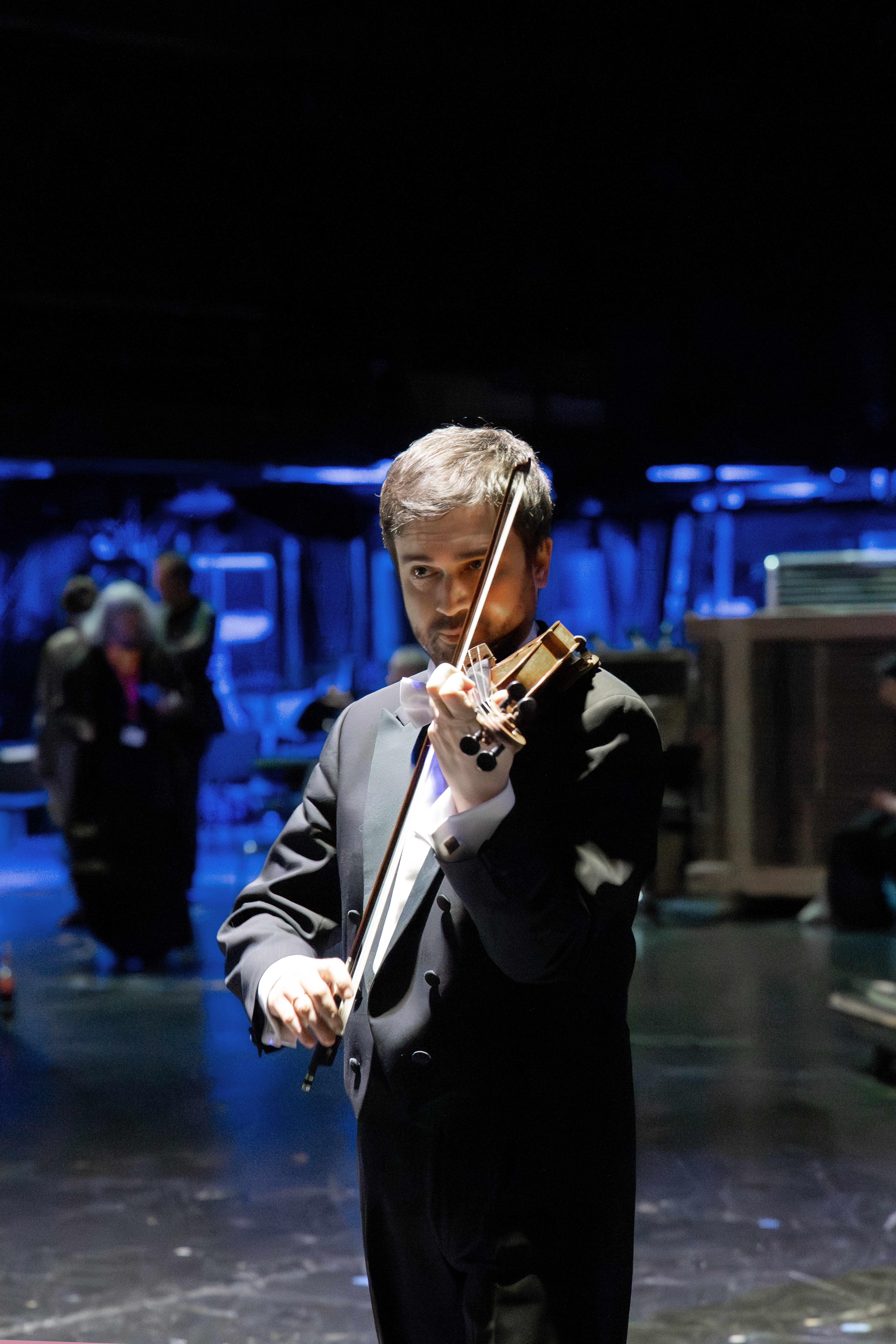 Ein Mann im Smoking spielt Geige auf einer schwach beleuchteten Bühne, auf der im unscharfen Hintergrund Menschen und Geräte zu sehen sind. Die blaue Beleuchtung schafft eine dramatische Atmosphäre.