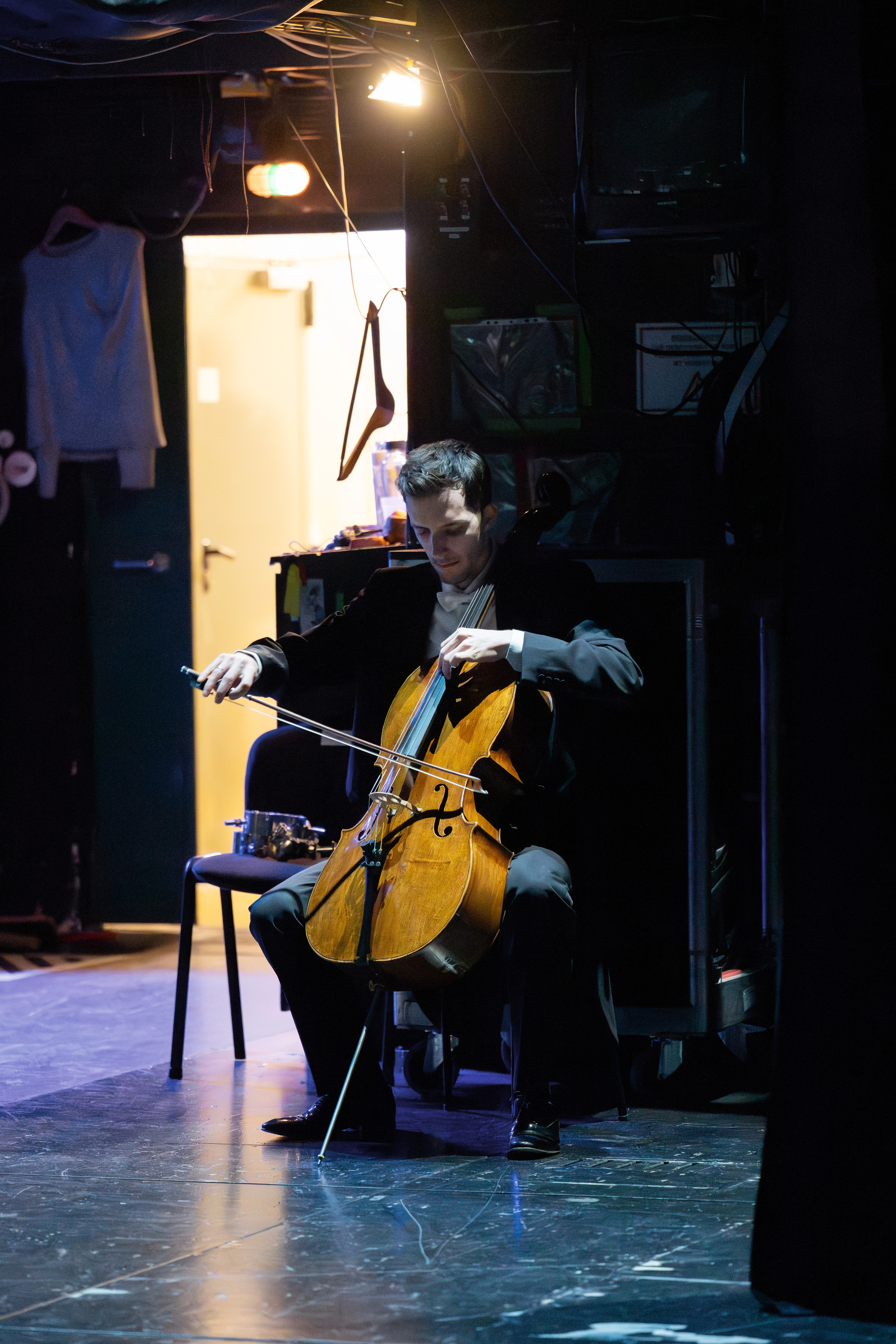 Ein Cellist in formeller Kleidung sitzt hinter der Bühne und spielt bei gedämpfter Beleuchtung Cello. Die Bühne ist dunkel, mit Kabeln über dem Kopf, einer offenen Tür und hängender Kleidung im Hintergrund.