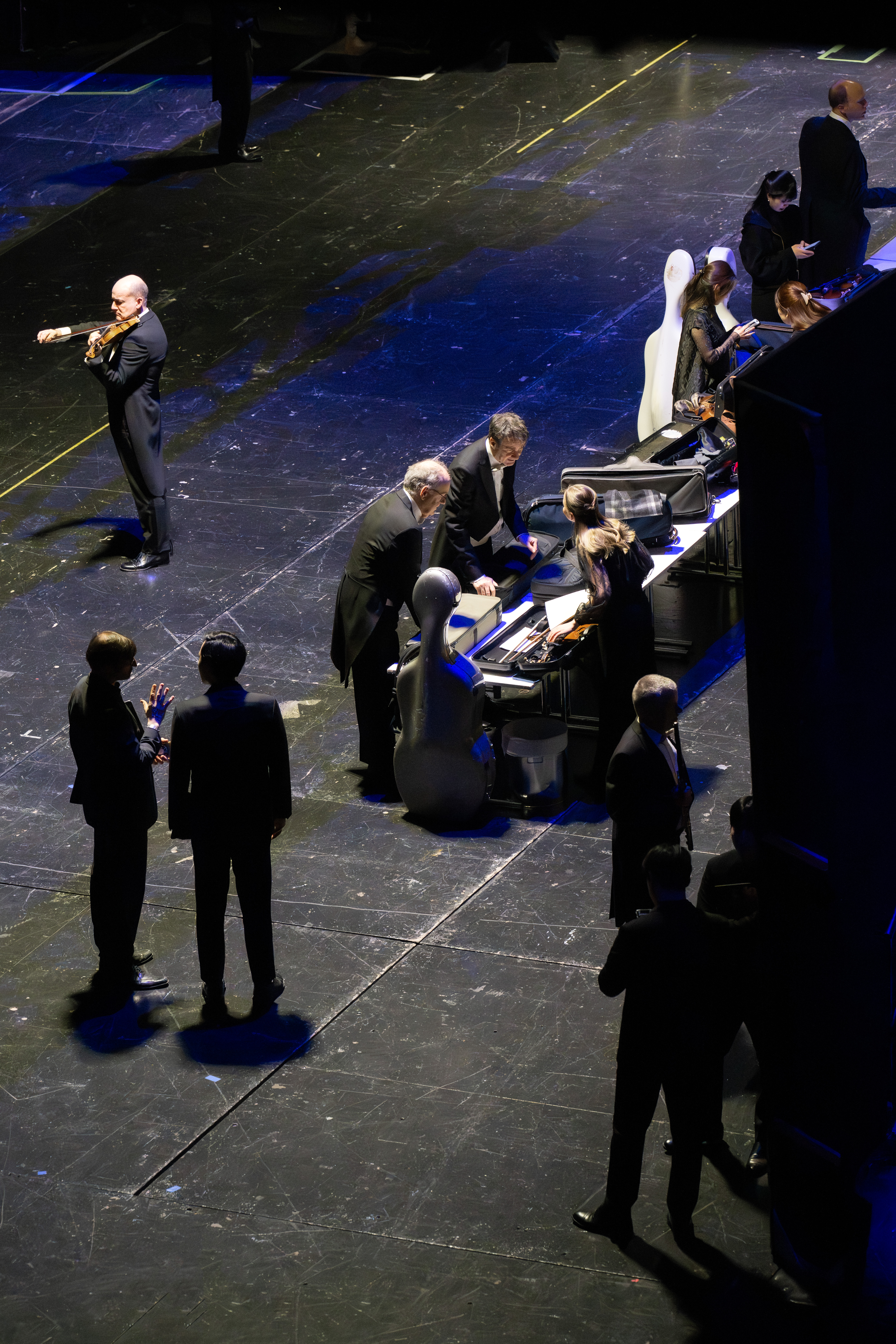 Musiker und Mitarbeiter versammeln sich auf einer schwach beleuchteten Bühne; eine Person spielt vorne auf einer Geige, während andere im Hintergrund an Musikinstrumenten und Geräten stehen oder arbeiten.