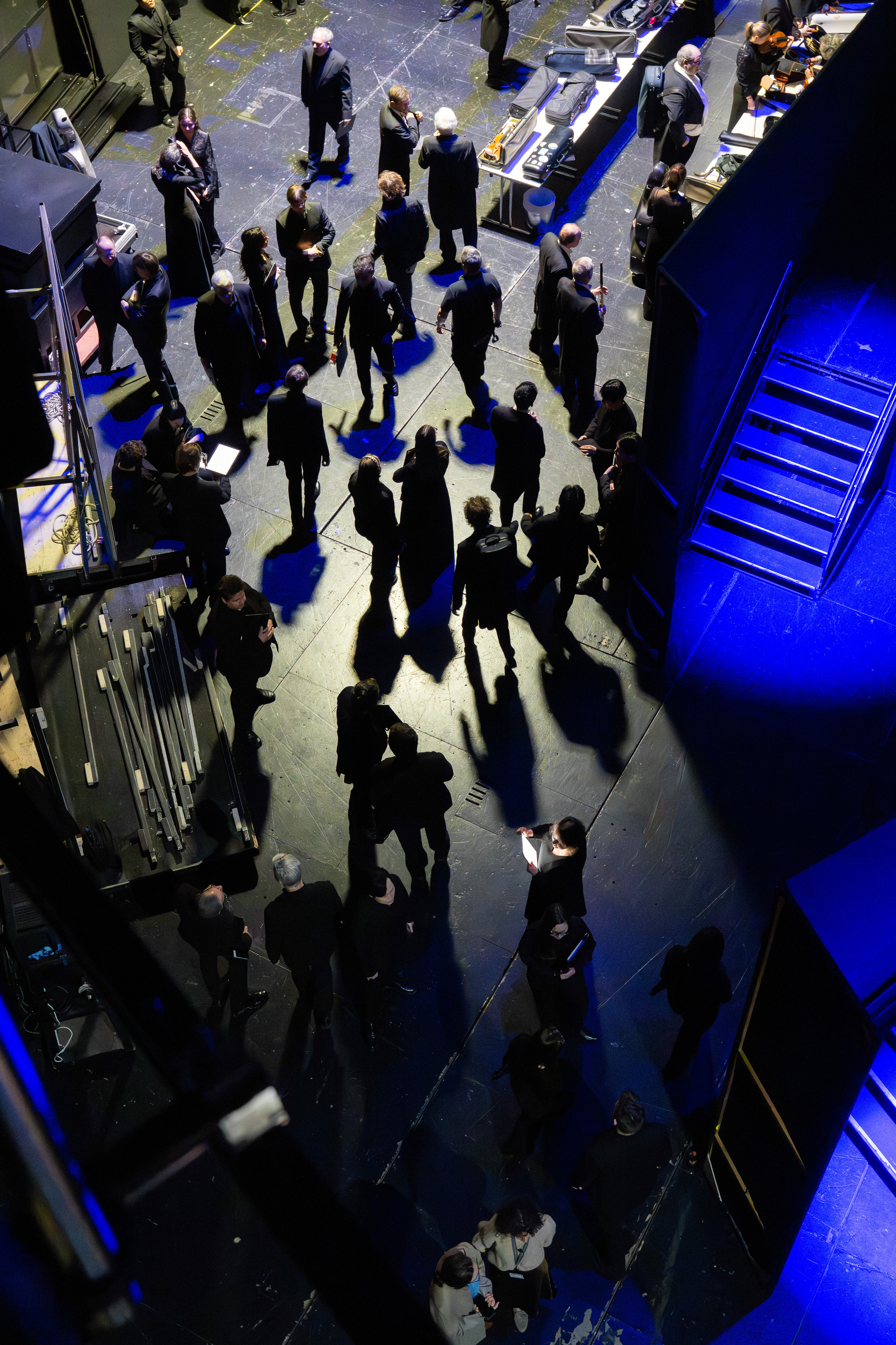 Eine Gruppe von Musikern in formeller schwarzer Kleidung versammelt sich um lange Tische hinter der Bühne, packt ihre Instrumente aus und bereitet sie für einen Auftritt vor. Die Szene wird von oben betrachtet.