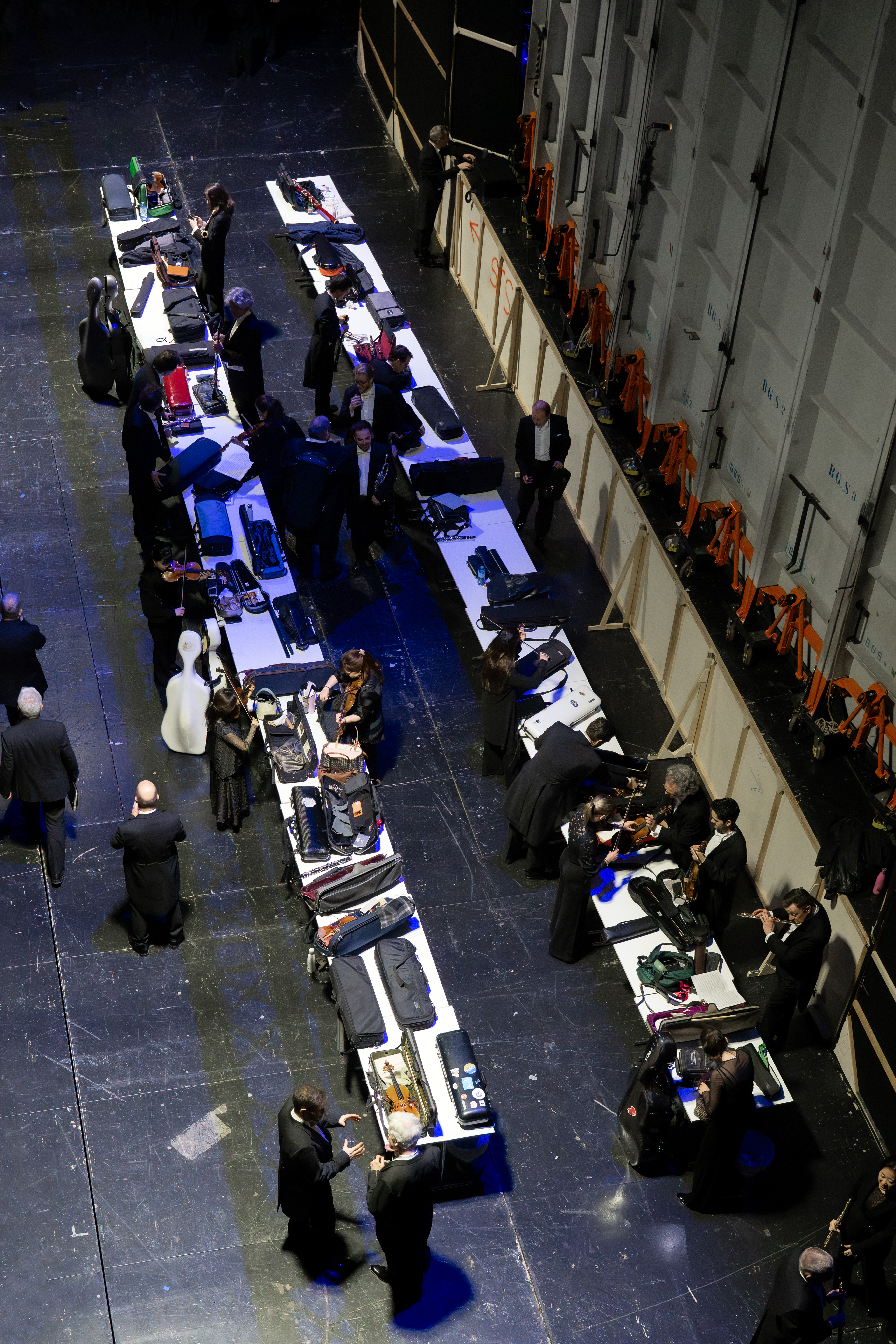 Eine Gruppe von Musikern in formeller schwarzer Kleidung versammelt sich um lange Tische hinter der Bühne, packt ihre Instrumente aus und bereitet sie für einen Auftritt vor. Die Szene wird von oben betrachtet.