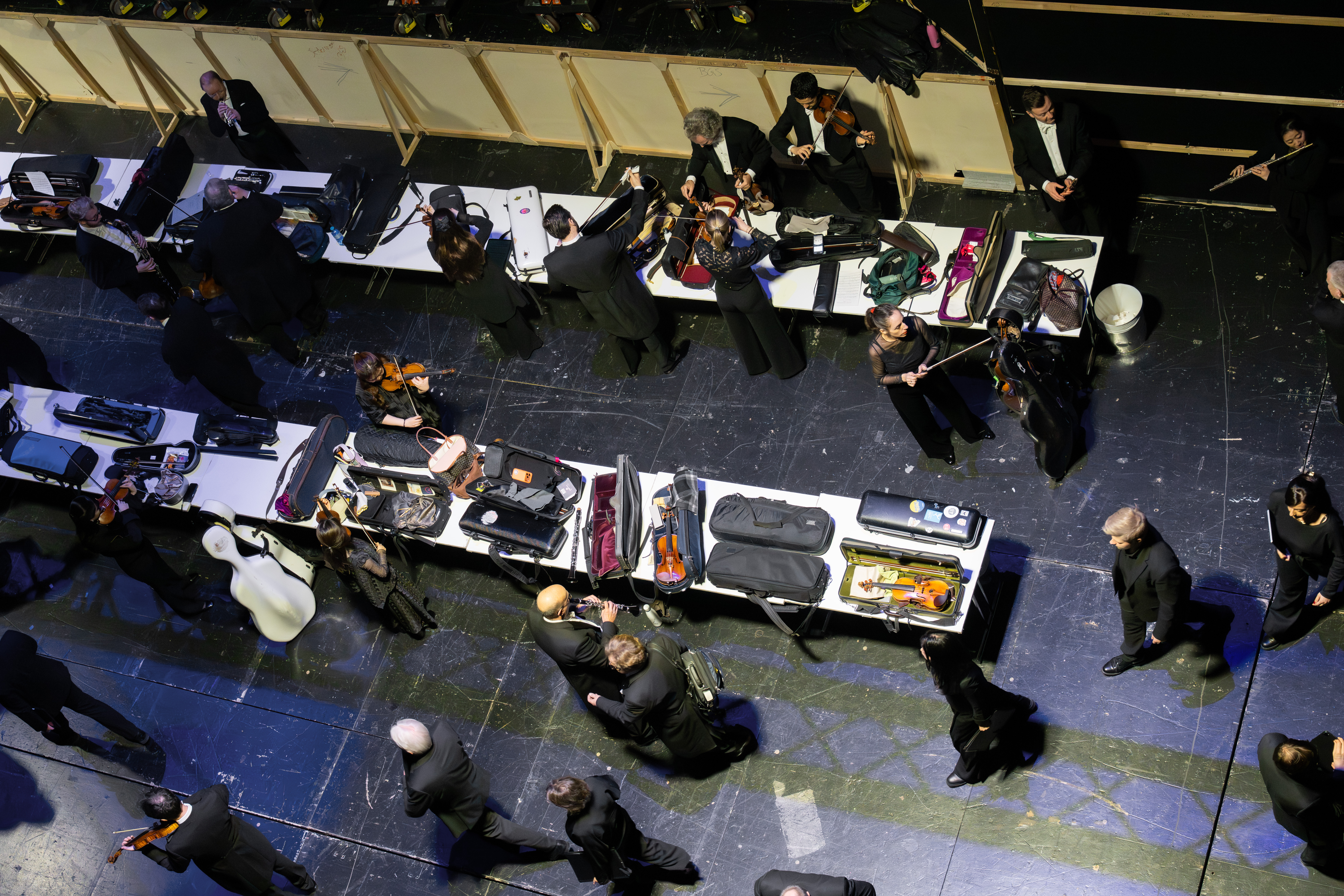 Eine Gruppe von Musikern in formeller schwarzer Kleidung versammelt sich um lange Tische hinter der Bühne, packt ihre Instrumente aus und bereitet sie für einen Auftritt vor. Die Szene wird von oben betrachtet.