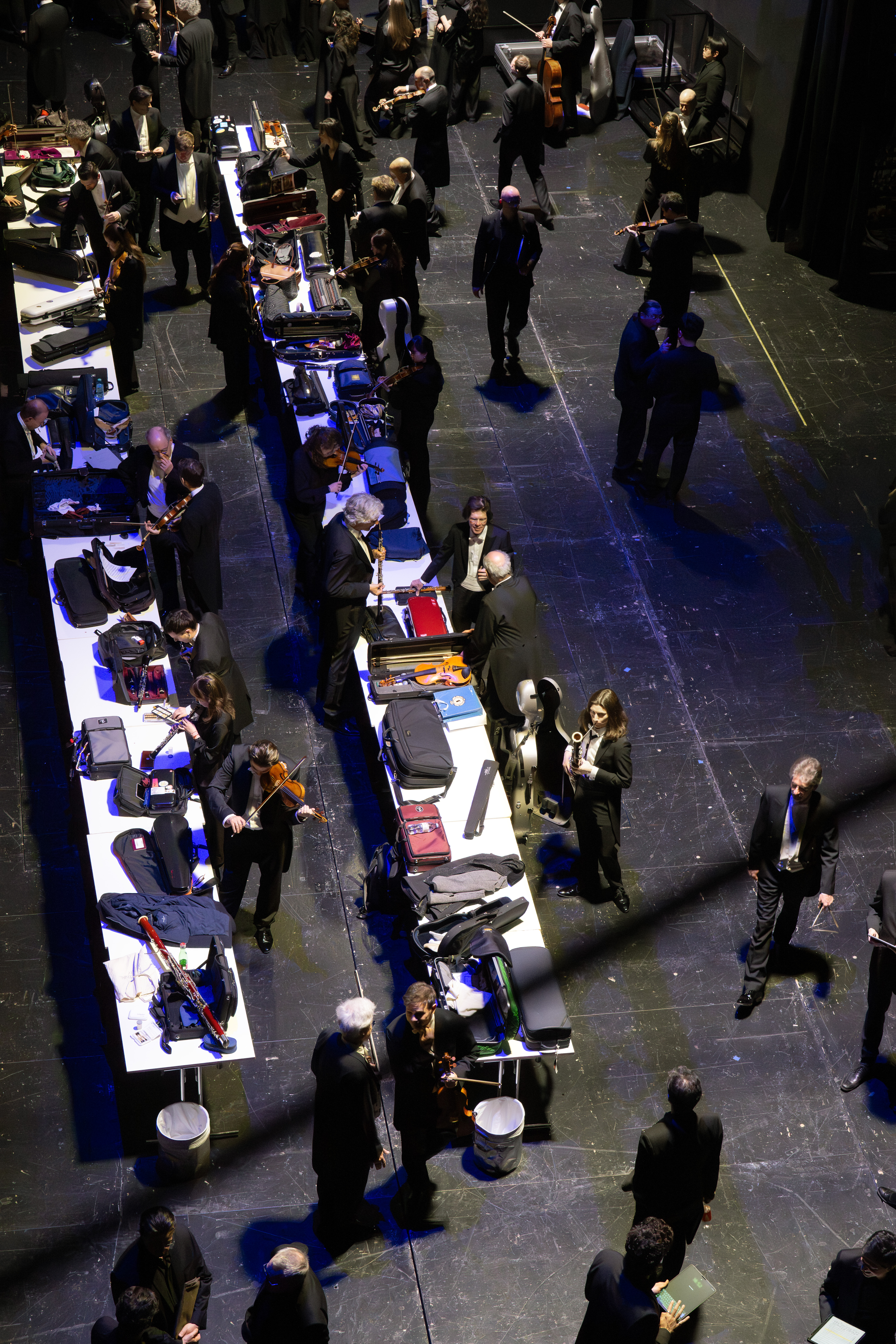 Eine Gruppe von Musikern in formeller schwarzer Kleidung versammelt sich um lange Tische hinter der Bühne, packt ihre Instrumente aus und bereitet sie für einen Auftritt vor. Die Szene wird von oben betrachtet.
