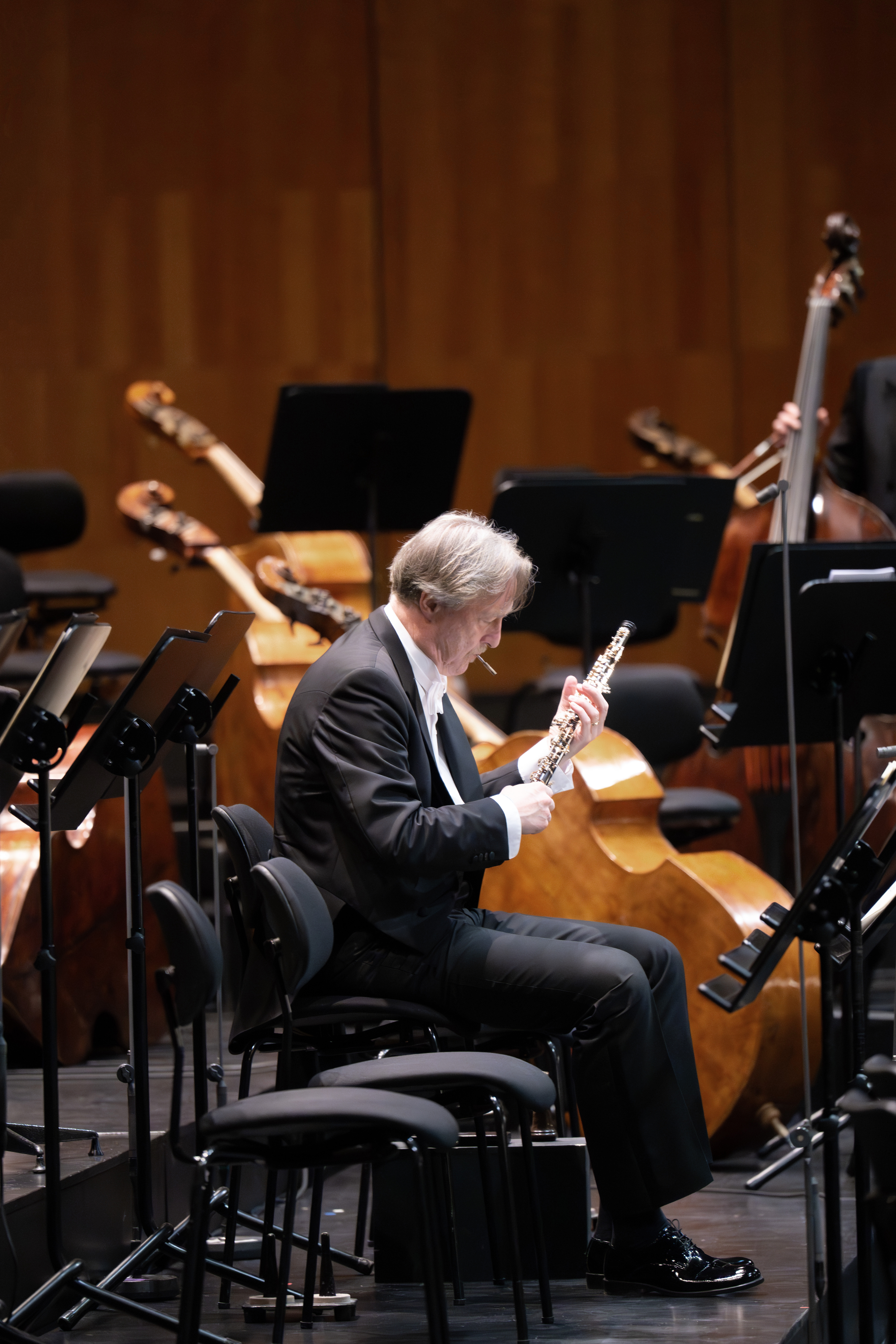 Ein Musiker in einem schwarzen Anzug sitzt auf der Bühne, hält eine Oboe in der Hand, umgeben von Stühlen und Streichinstrumenten, und bereitet sich auf einen Orchesterauftritt vor.