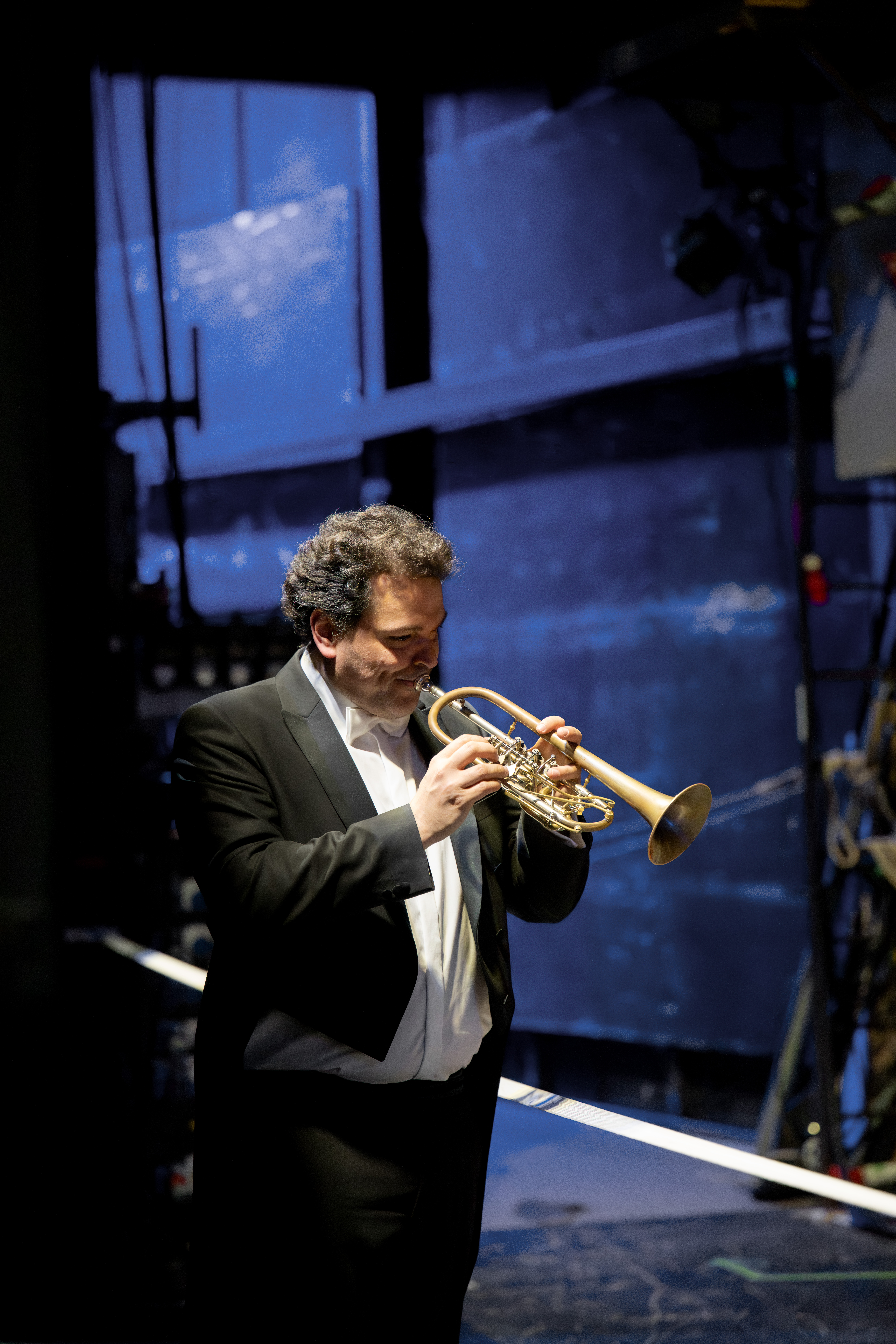 Ein Mann im schwarzen Smoking spielt hinter der Bühne Trompete, er steht in der Nähe von Bühnenausrüstung und Kabeln, im Hintergrund ist eine blau beleuchtete Wand zu sehen.