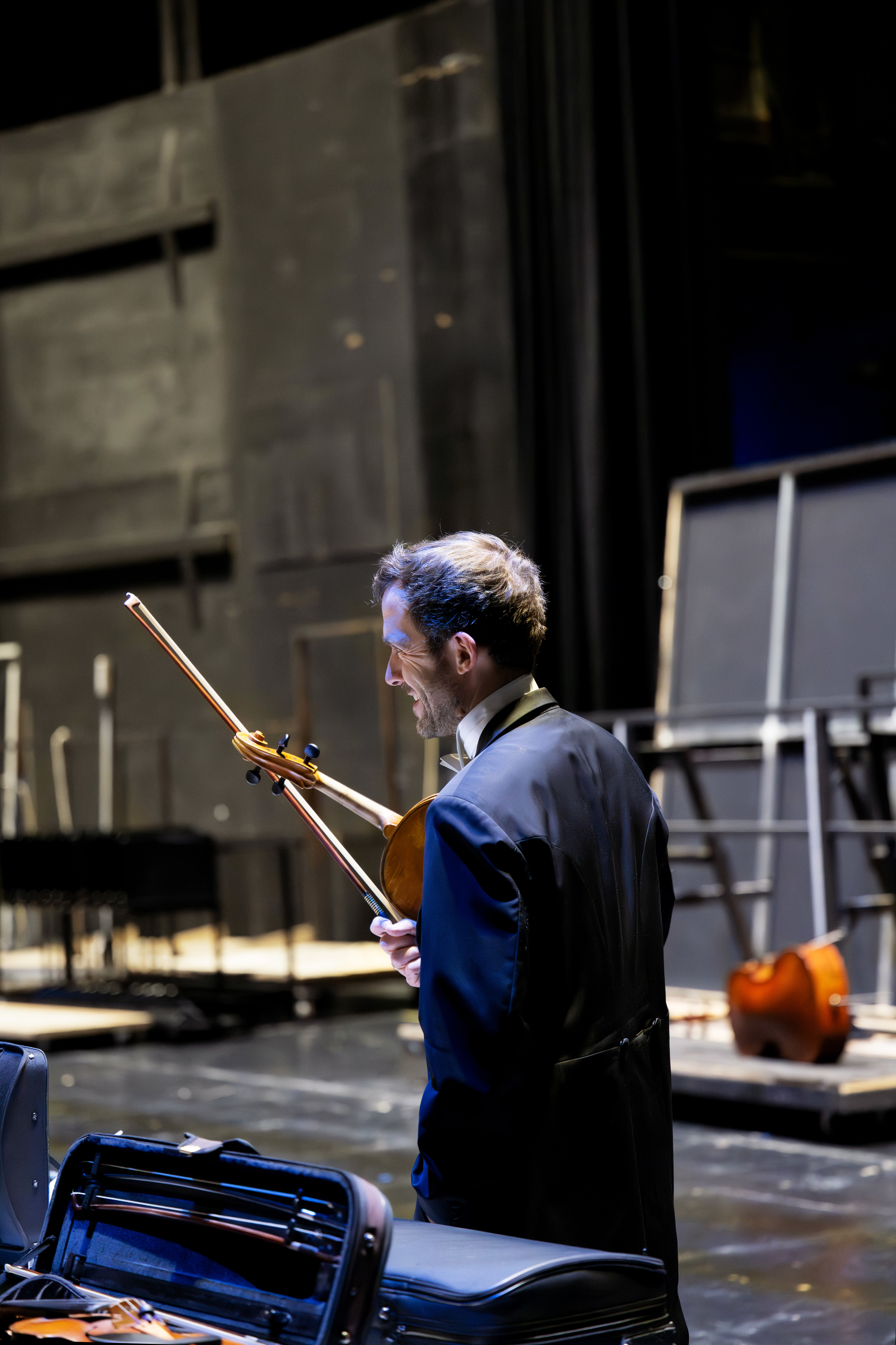 Ein Mann im Smoking hält eine Bratsche und einen Bogen hinter der Bühne eines schwach beleuchteten Theaters. Er steht in der Nähe von Musikkoffern und Instrumenten, während im Hintergrund Bühnenausrüstung und Requisiten zu sehen sind.