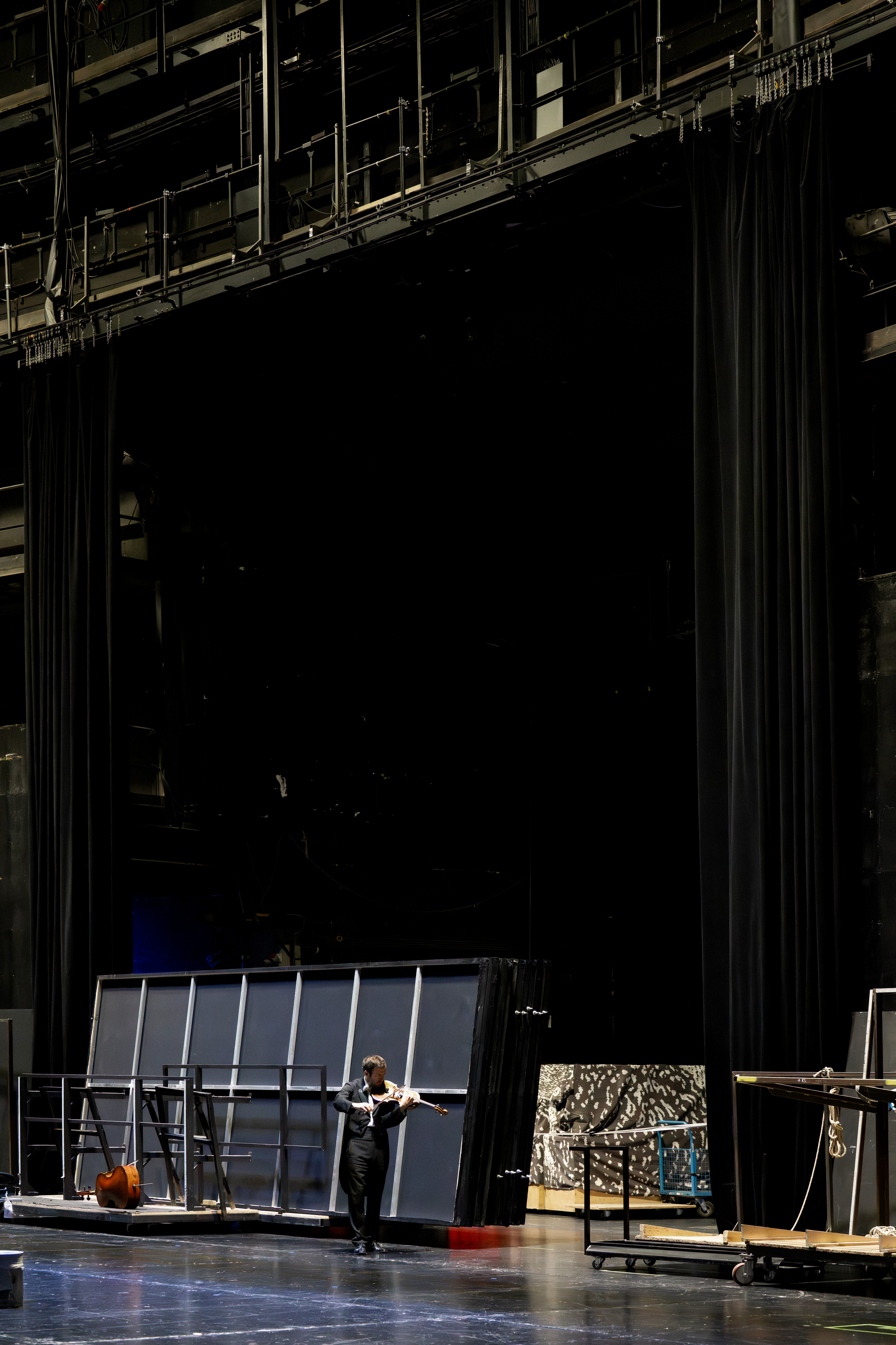 Eine schwarz gekleidete Person arbeitet hinter der Bühne in der Nähe großer Kulissen, schwarzer Vorhänge und verschiedener Geräte auf einer schwach beleuchteten Theaterbühne.