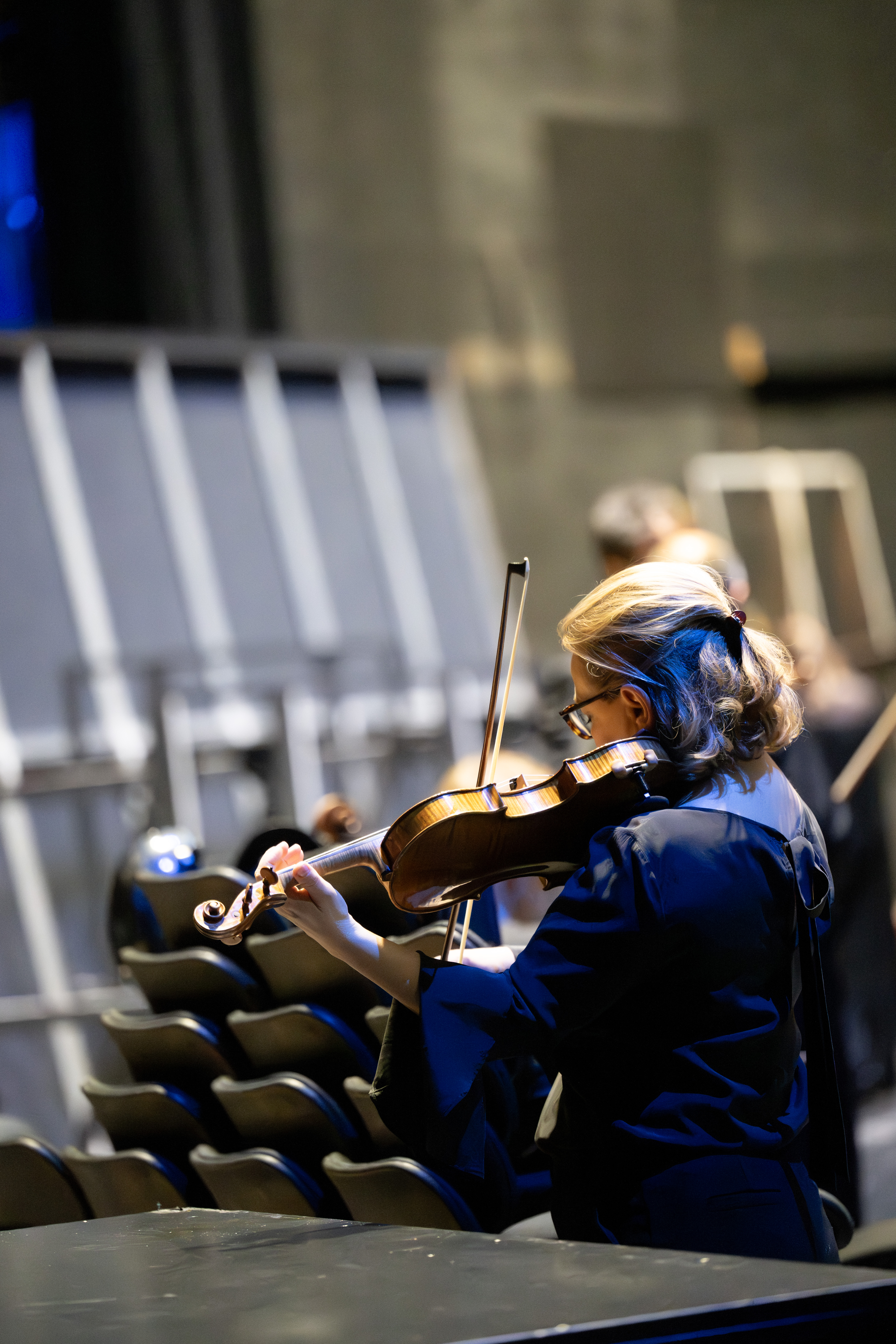 Eine Frau in einem dunklen Kleid spielt auf der Bühne, vom Publikum abgewandt, Geige. Im Hintergrund sind leere schwarze Stühle und verschwommene Figuren zu sehen.