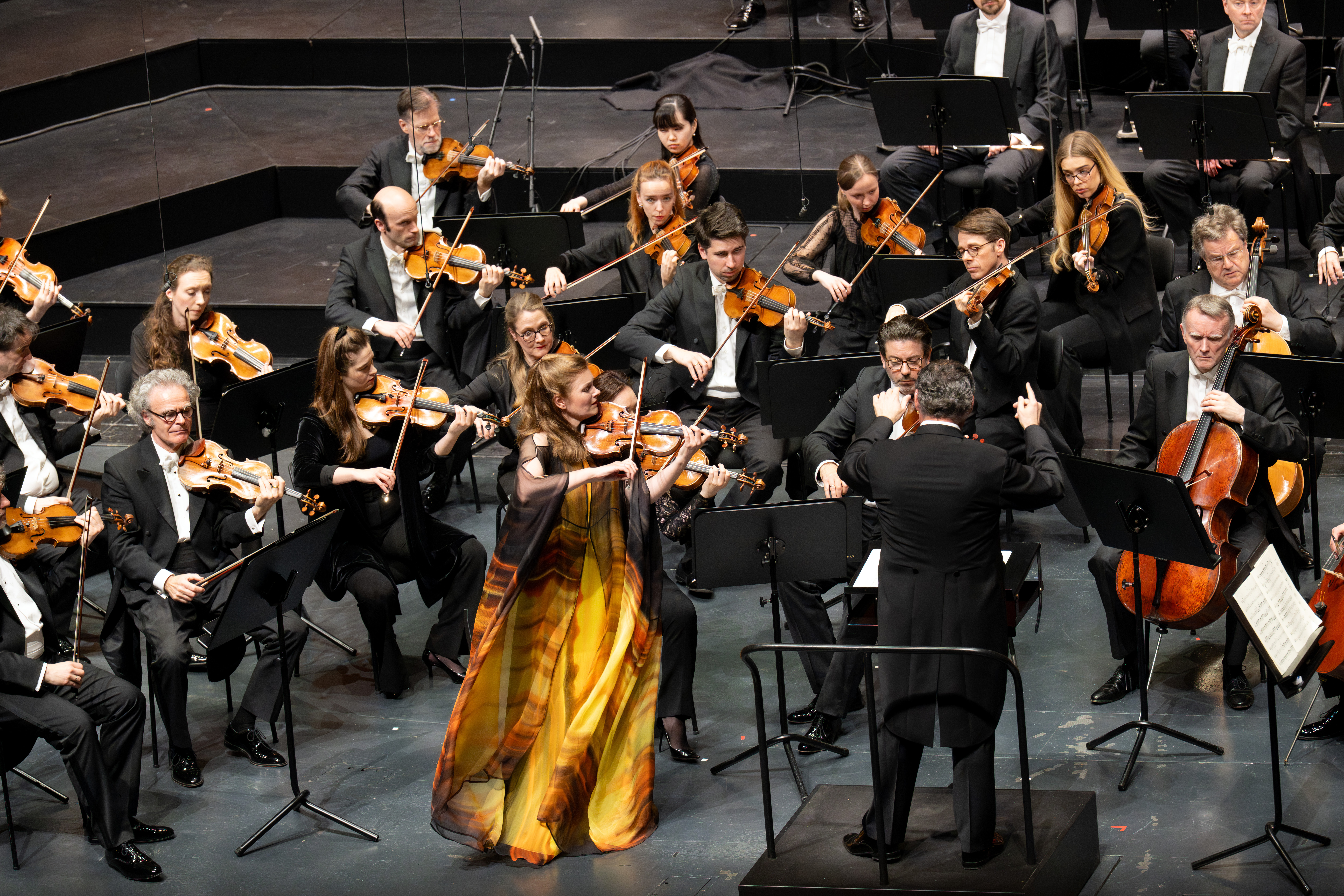 Eine Frau in einem wallenden gelb-orangefarbenen Kleid spielt ein Geigensolo mit einem Orchester auf der Bühne, während ein Dirigent in einem schwarzen Anzug die Musiker leitet, die ebenfalls formell gekleidet sind und Streichinstrumente spielen.