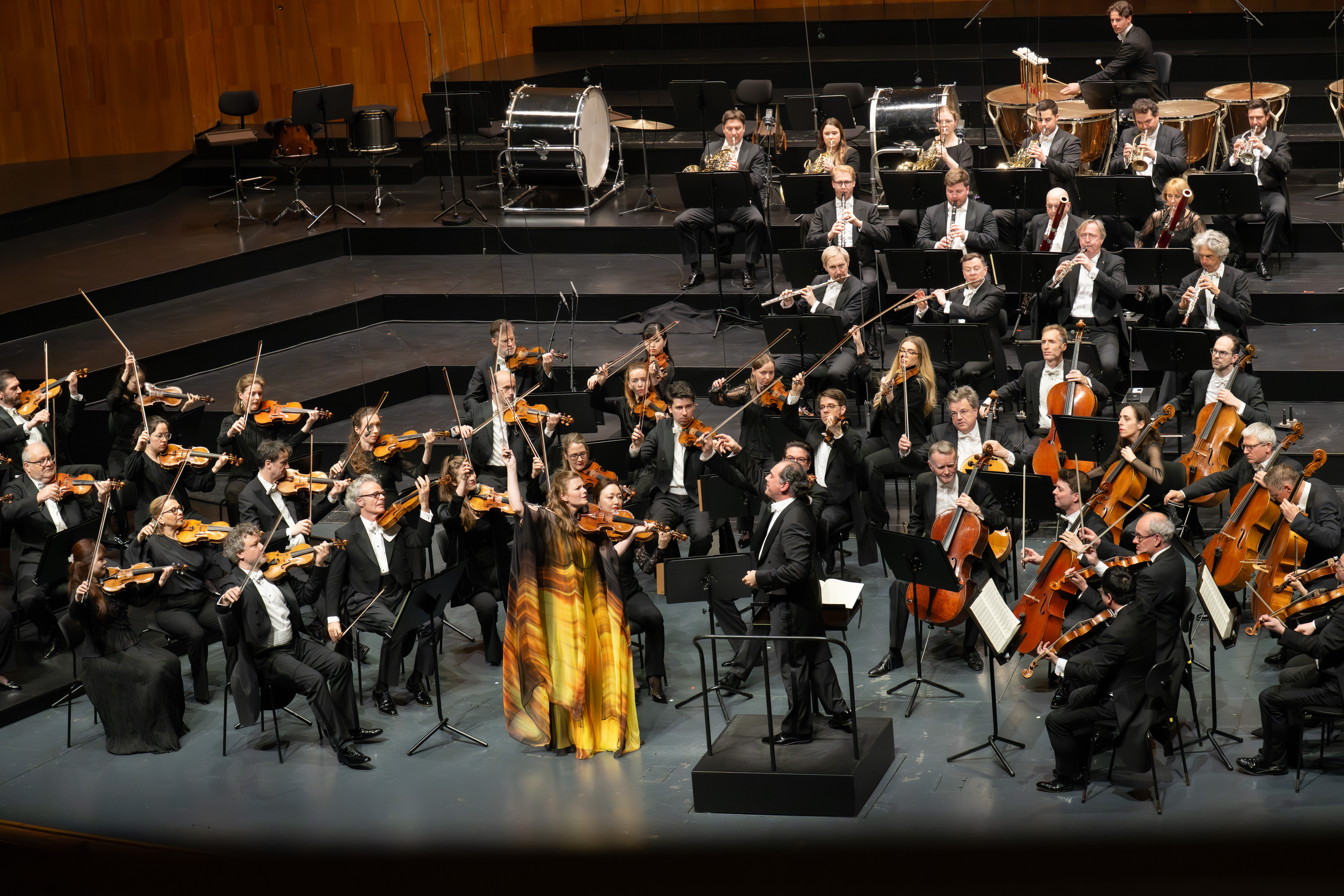 Ein Symphonieorchester spielt auf der Bühne; die Musiker spielen Streicher, Holz- und Blechblasinstrumente. Ein Dirigent leitet sie, während ein Solist in einem gelben Kleid in der Mitte steht.