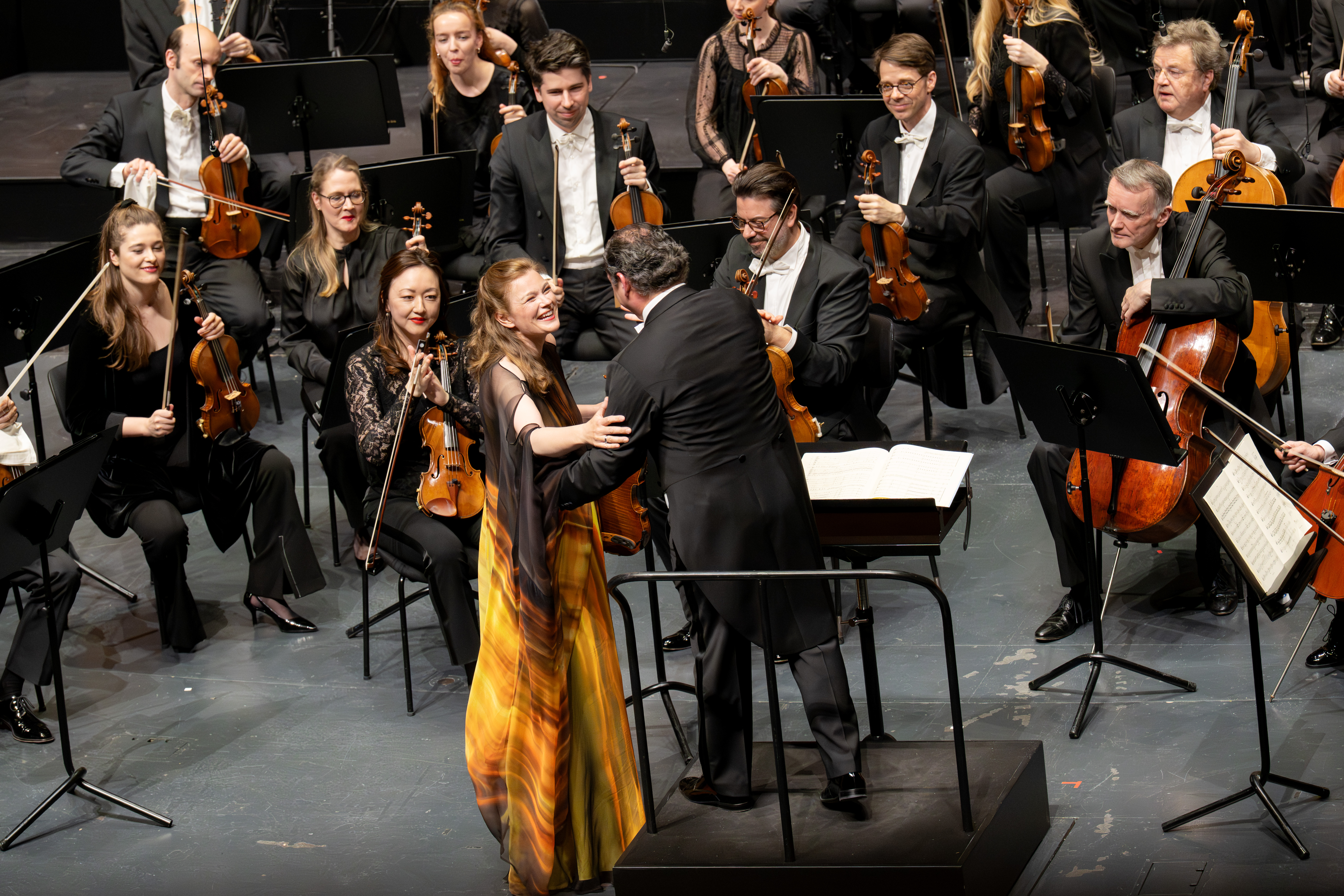 Eine lächelnde Solistin in einem gelben Kleid hält mit einem Dirigenten auf der Bühne Händchen, umgeben von sitzenden Orchestermusikern mit Streichinstrumenten, die alle engagiert und fröhlich wirken.