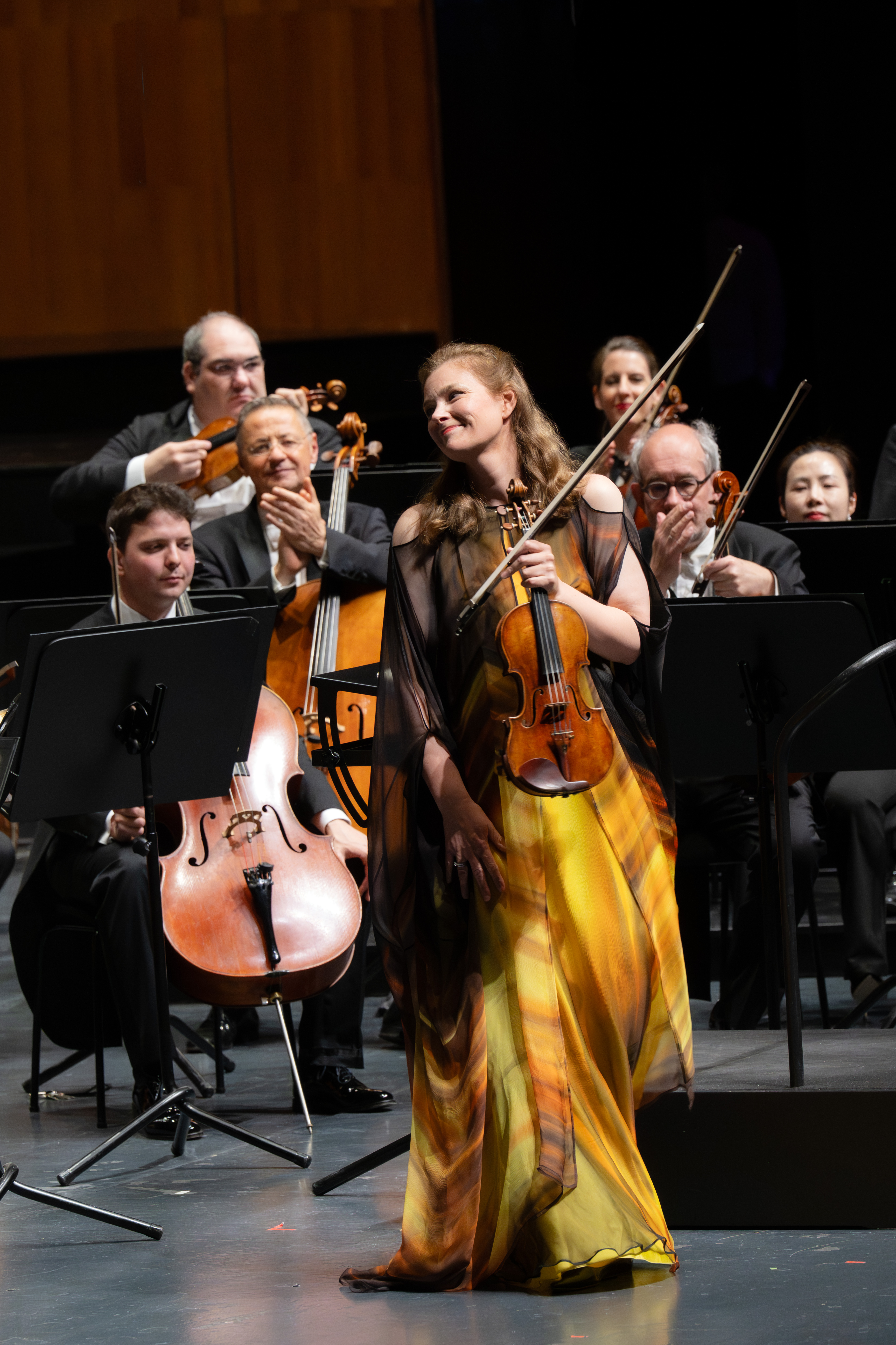 Eine lächelnde Geigerin in einem gelb-braunen Kleid steht vor einem sitzenden Orchester auf der Bühne. Einige Musiker klatschen und schauen ihr zu, während Notenständer und Instrumente sie umgeben.