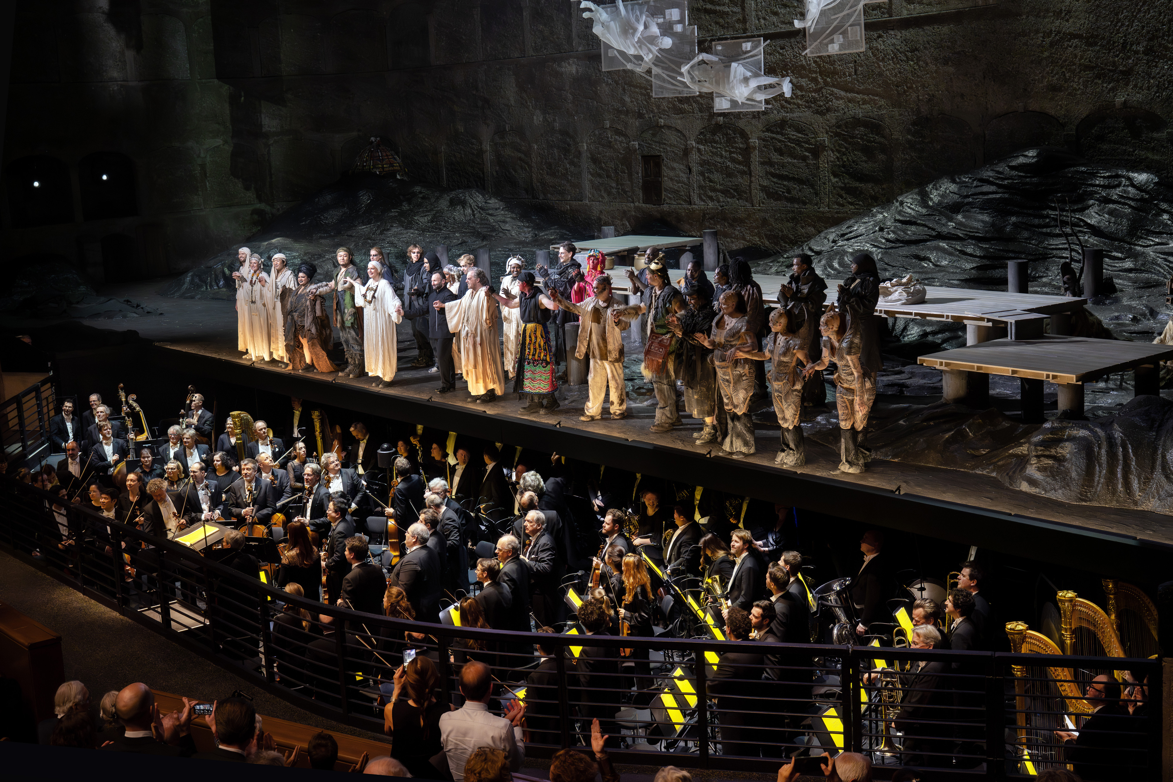 A group of performers in costume take a bow on a rocky stage, while an orchestra plays below. The audience watches from the foreground in a large, dramatic theater setting.