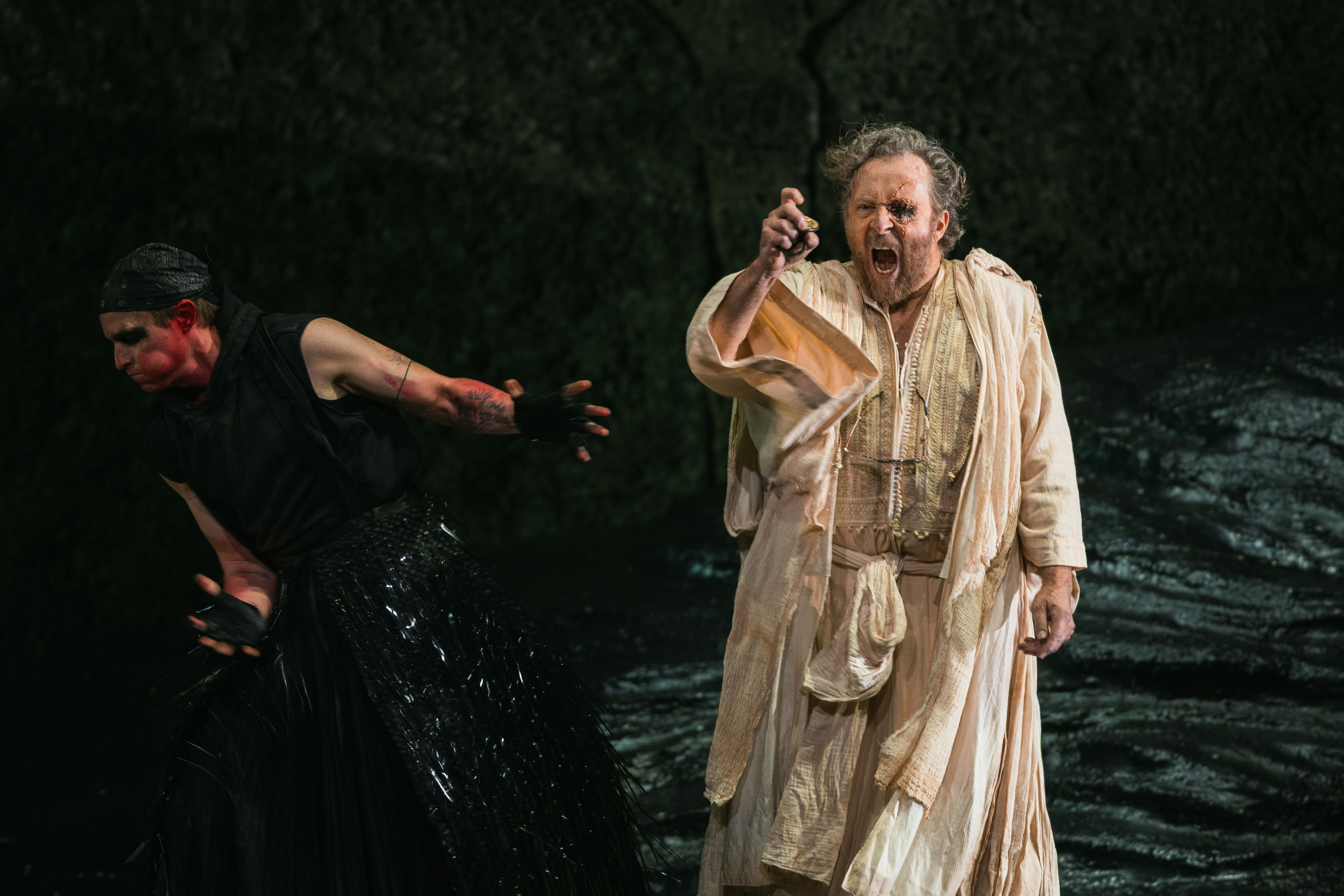 Zwei Schauspieler treten auf der Bühne auf; der eine in einem dunklen, dramatischen Kostüm mit Gesichtsbemalung schreckt zurück, während der andere in einem hellen, fließenden Gewand schreit und kraftvoll gestikuliert und in einer angespannten Szene ein Tuch hält.