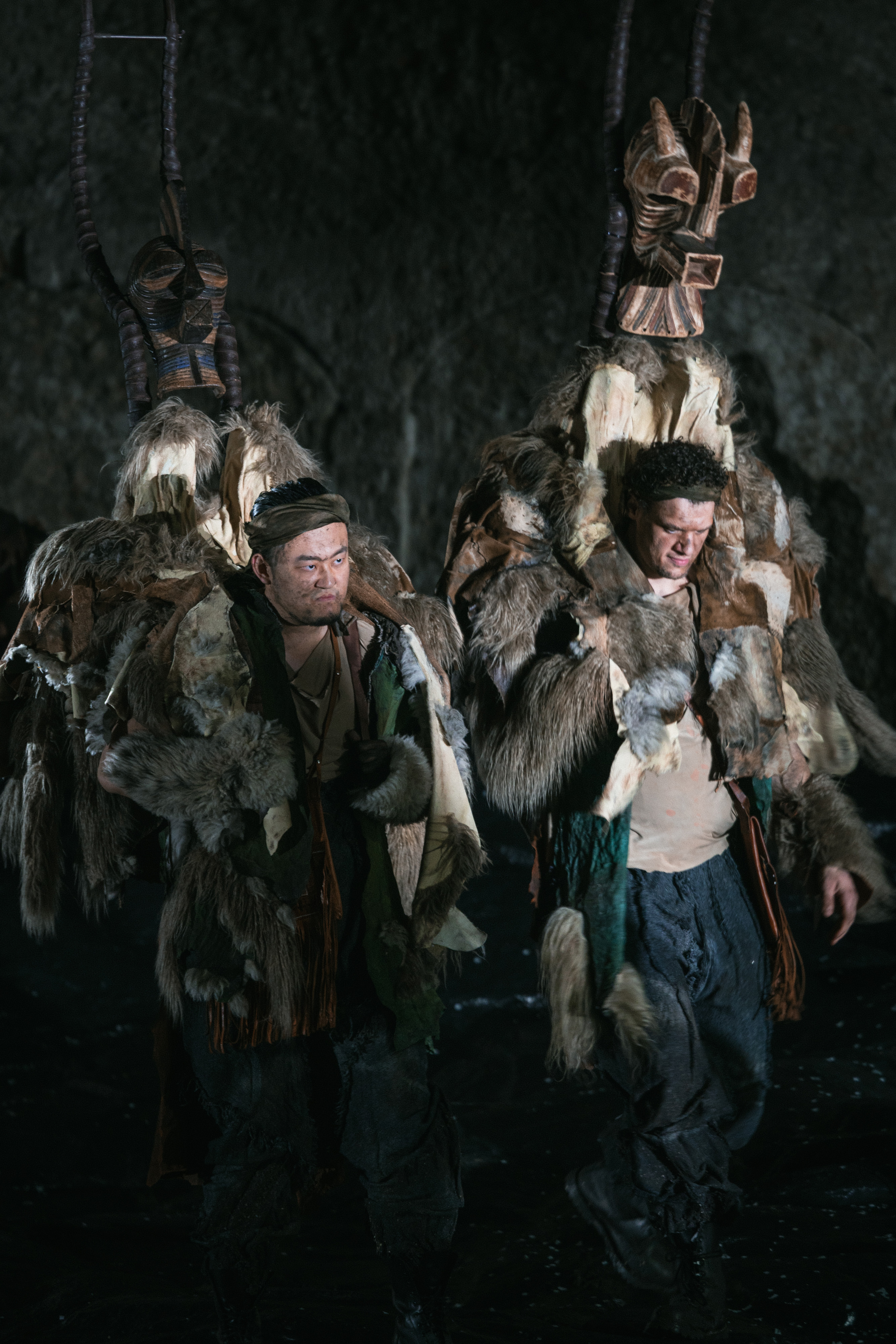 Two people dressed in fur garments and elaborate headdresses with animal-like masks walk in a dimly lit, rocky environment, suggesting a ritual or performance.