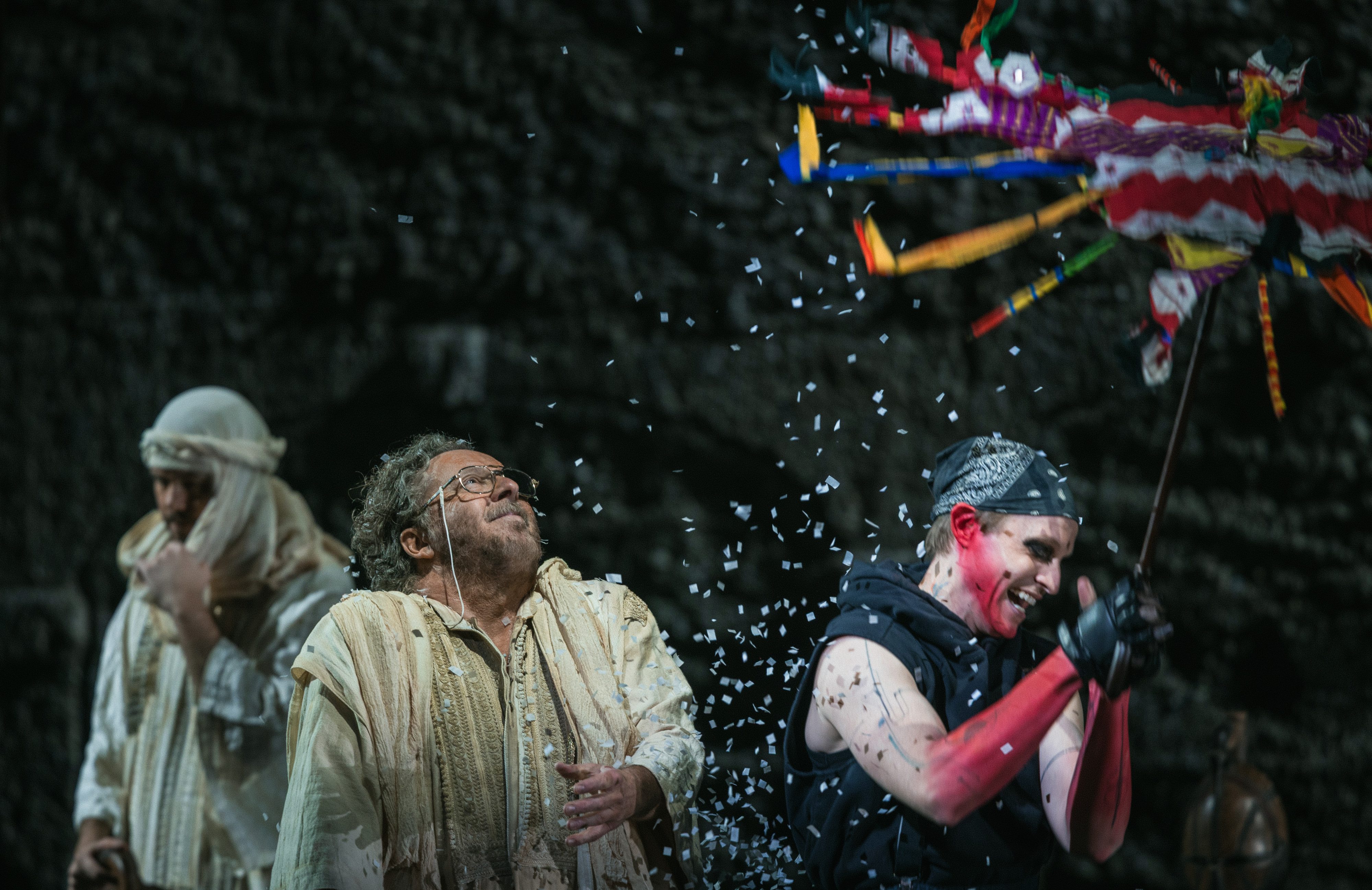 Three people on stage celebrate as one, dressed in black with pink sleeves, breaks a colorful piñata, showering confetti. Another person in beige looks up, smiling, while confetti falls around them.