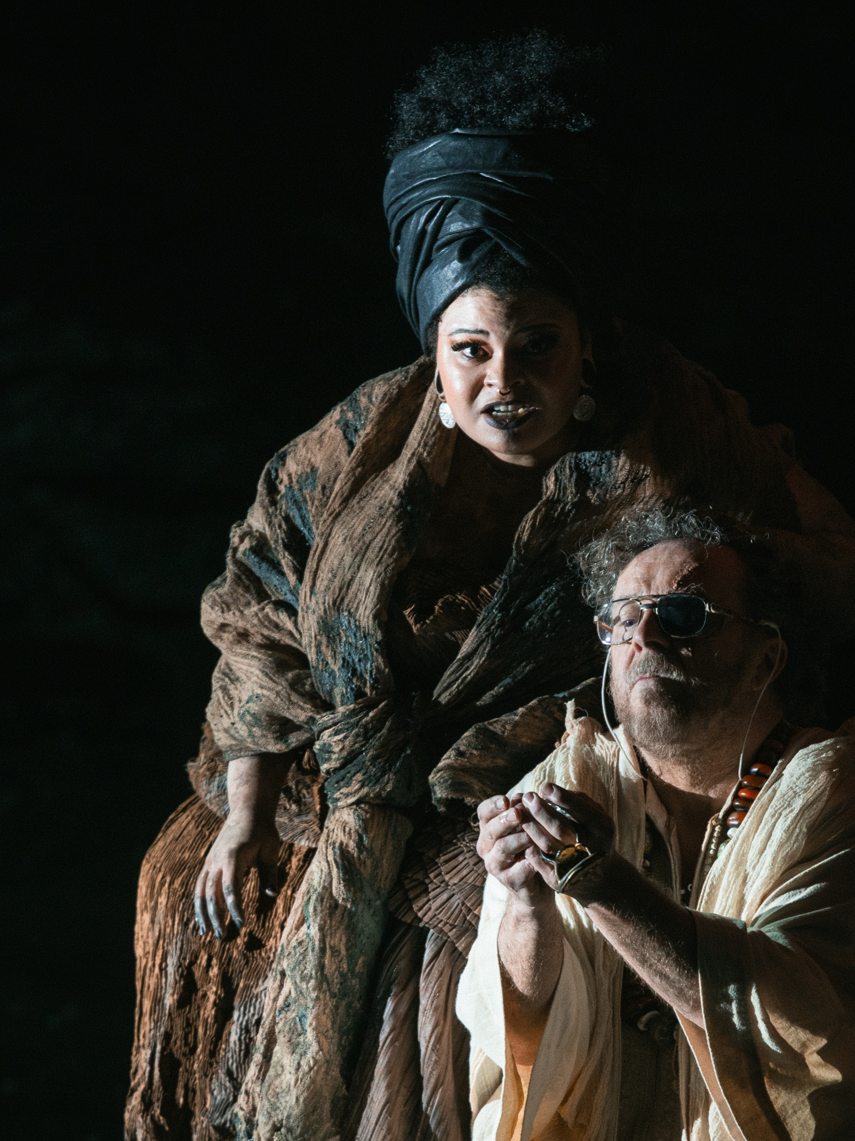 A woman in a textured brown robe and black headwrap stands behind a seated man with an eyepatch and beads, who is holding an object in his hands. They are both dramatically lit against a dark background.